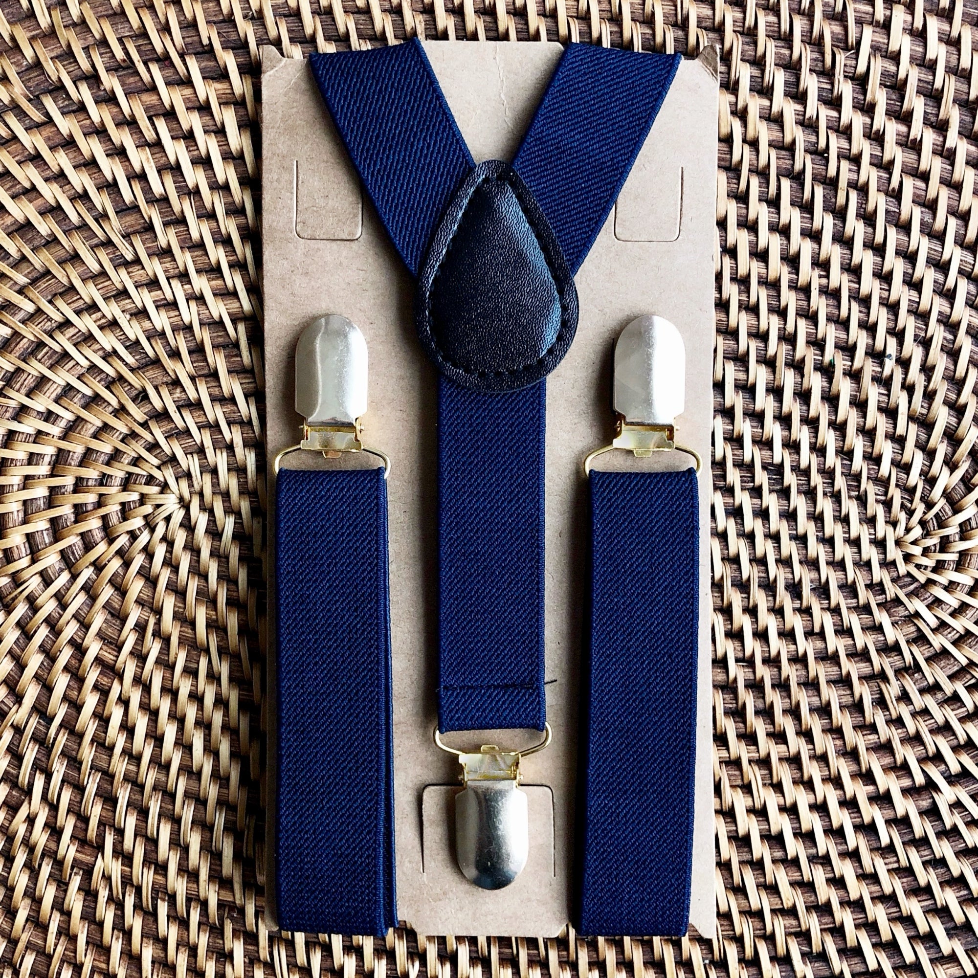 Navy blue suspenders with silver clips on a cardboard backing against a woven background