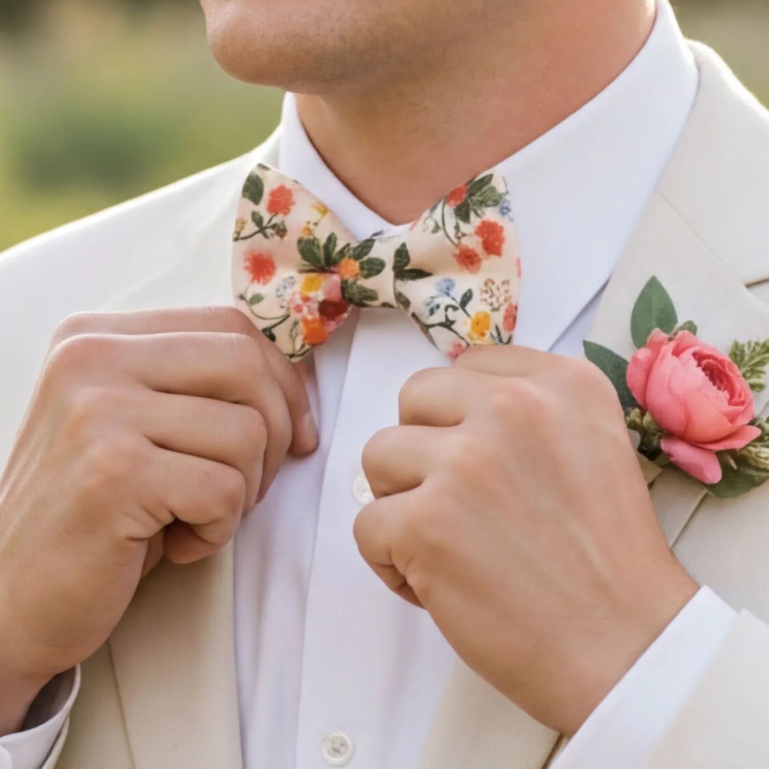 A man in a white suit and floral bow tie is adjusting his bow tie, with a pink rose boutonniere pinned to his lapel.