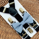 Dusty blue bow tie with bronze clips on black suspenders for groomsmen, ring bearer outfit and wedding on a woven surface.
