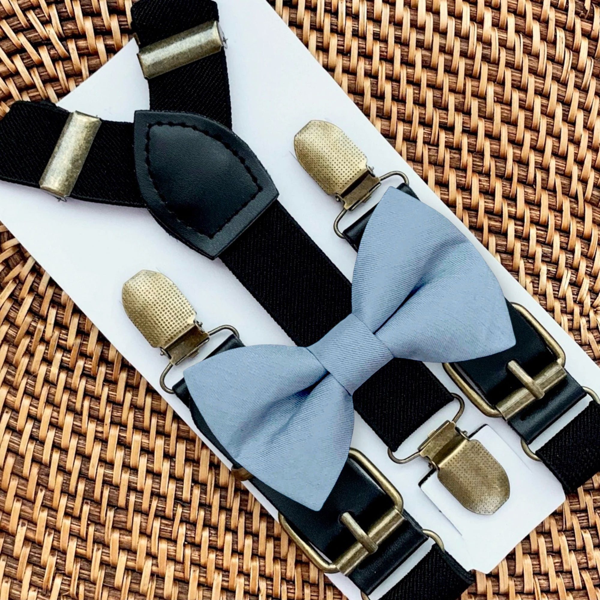 Dusty blue bow tie with bronze clips on black suspenders for groomsmen, ring bearer outfit and wedding on a woven surface.