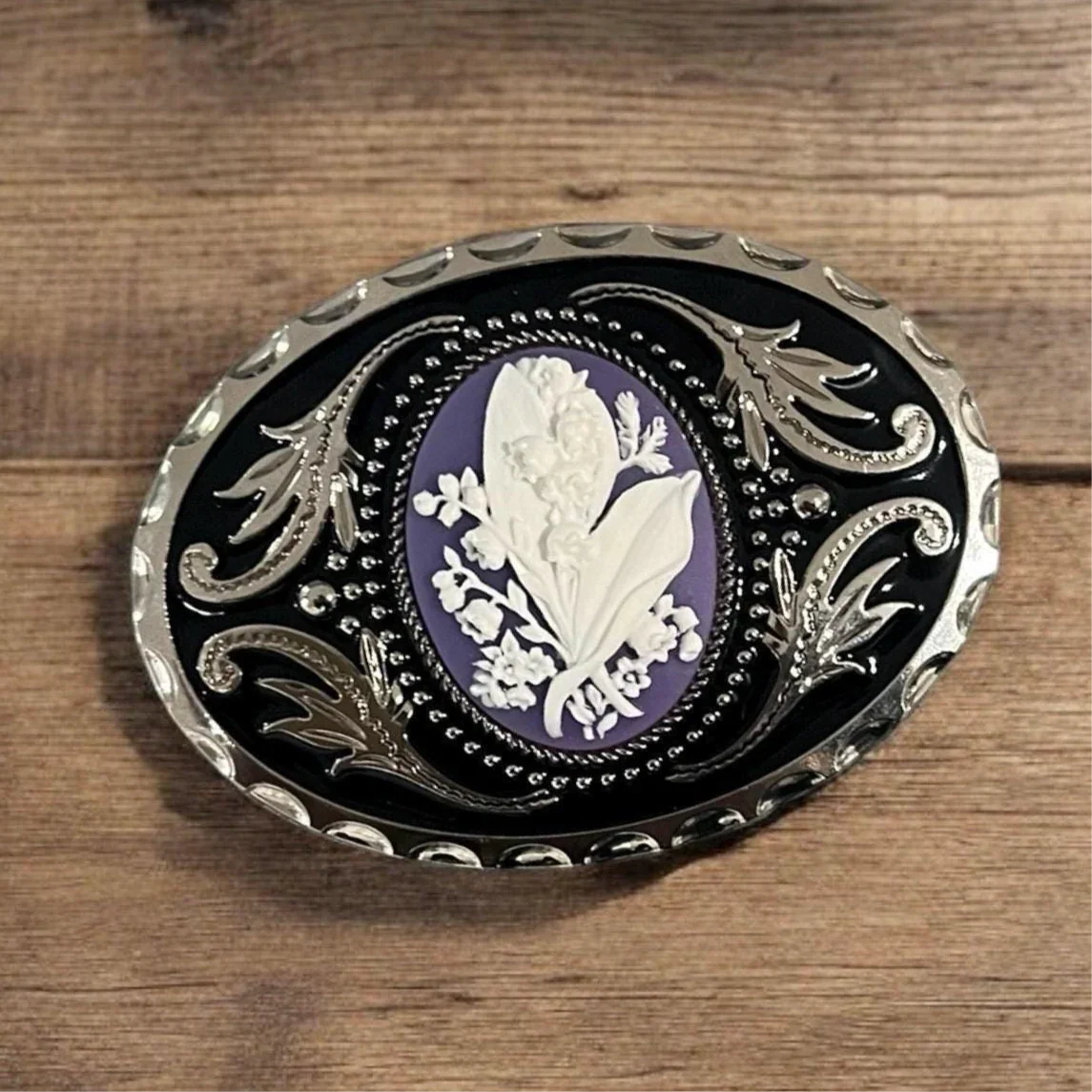 Oval belt buckle with silver scrollwork on black enamel and a purple center featuring white lily of the valley relief