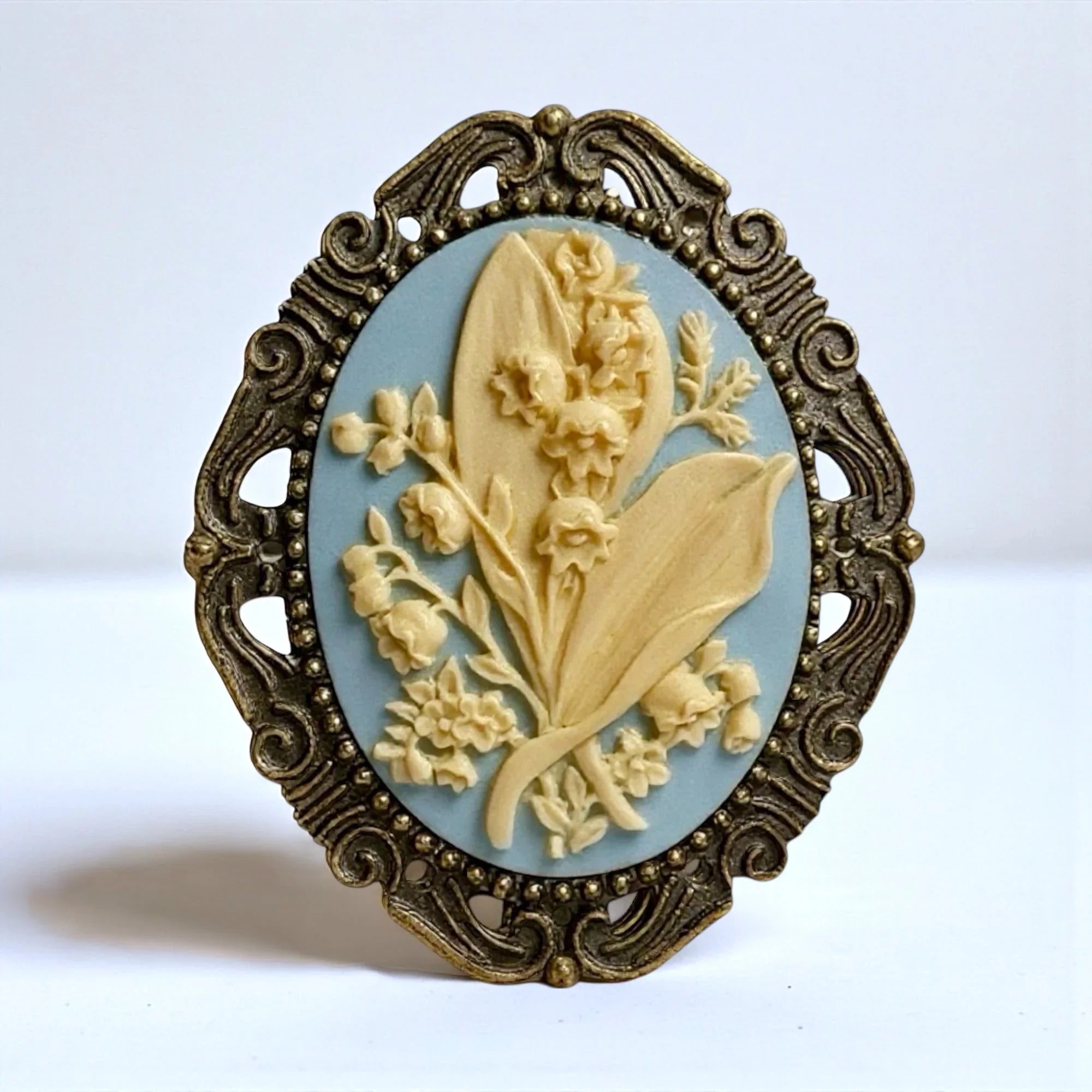 May flower Lily of the Valley brooch with blue and ivory floral design and ornate frame on a white background