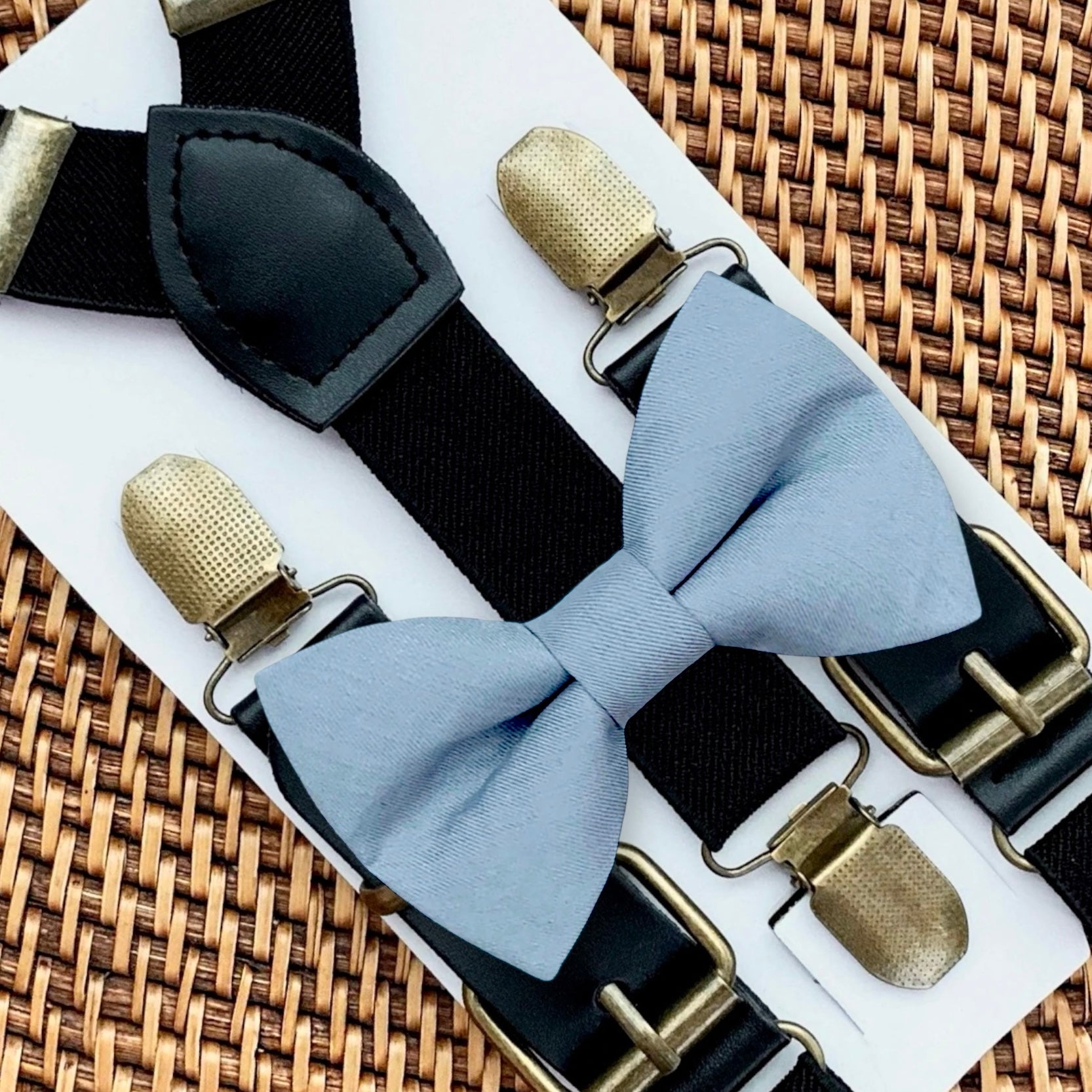 Dusty blue bow tie with bronze clips on black suspenders for groomsmen, ring bearer outfit and wedding on a woven surface