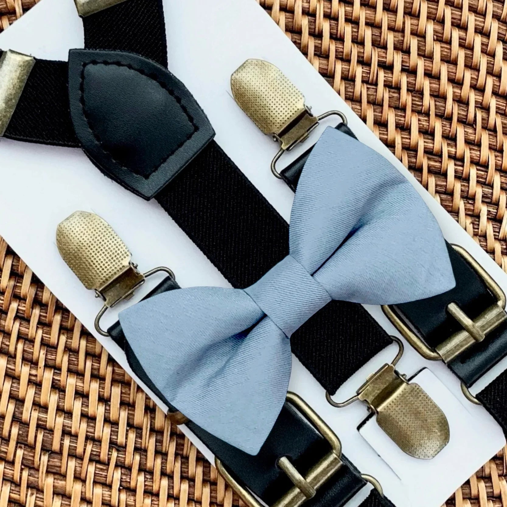 Dusty blue bow tie with bronze clips on black suspenders for groomsmen, ring bearer outfit and wedding on a woven surface