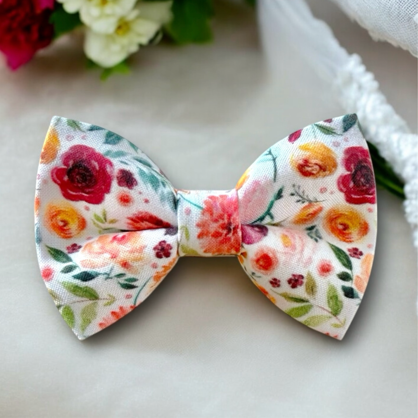 A pre-tied bow tie with a floral pattern, predominantly featuring red and yellow flowers, on a light background, displayed on a white surface with floral decorations in the background.