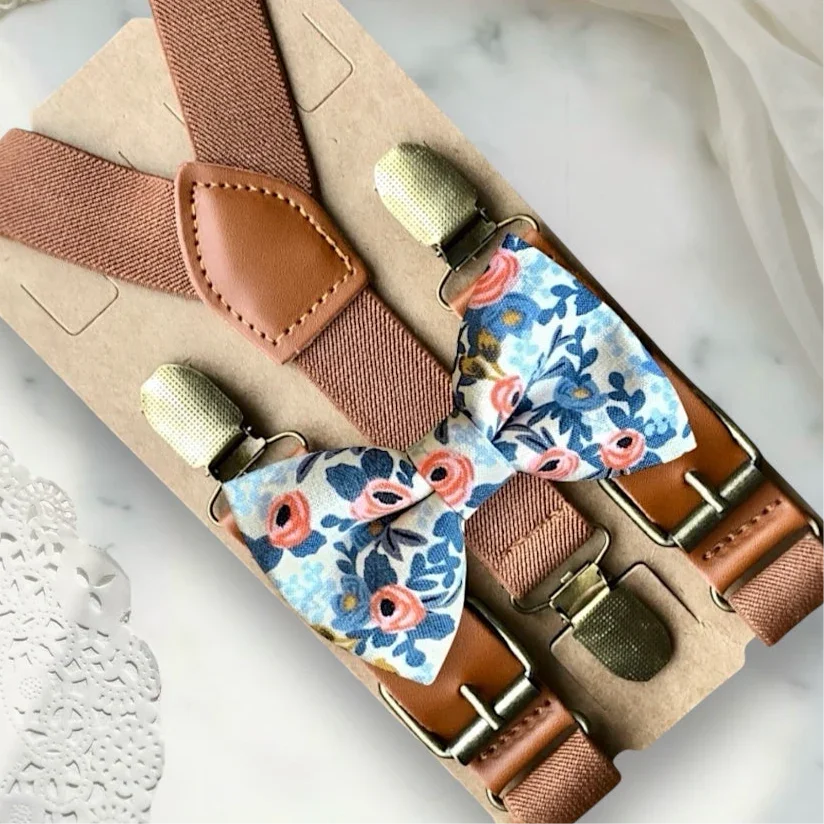 Angled view of white floral bow tie with cognac brown leather suspenders on a woven wicker surface