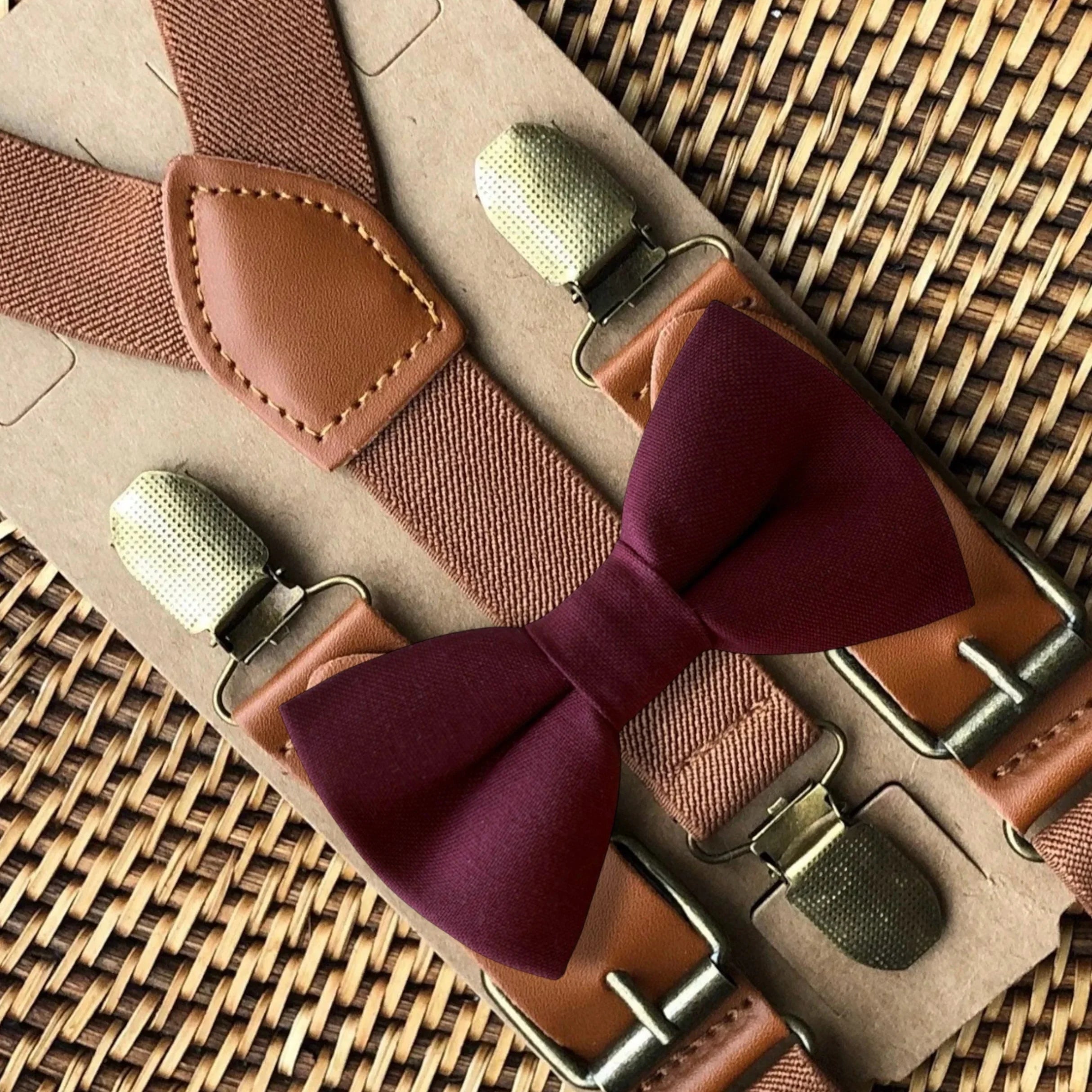 Cabernet burgundy bow tie with cognac buckle suspenders laid on a light surface.