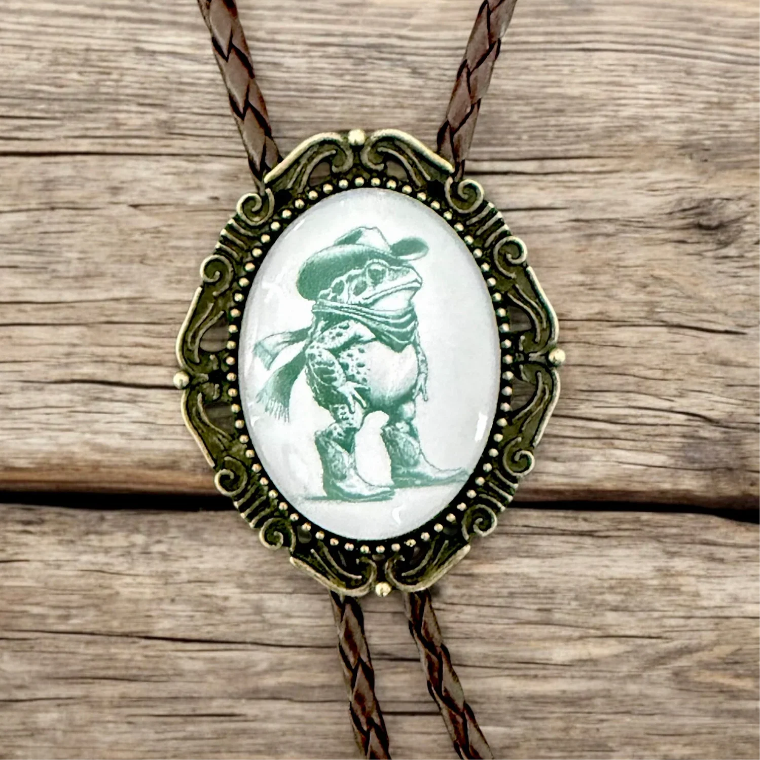 Bolo tie with a frog wearing a cowboy hat on an oval pendant, set against a wooden background.