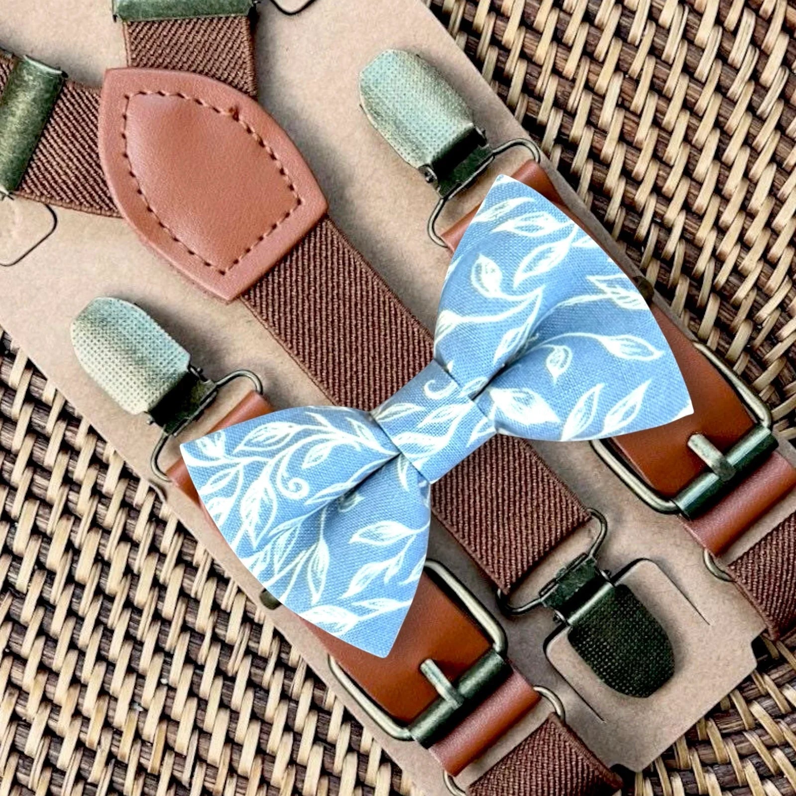 Dusty blue bow tie with white floral accents and dark brown suspenders on a beige display board