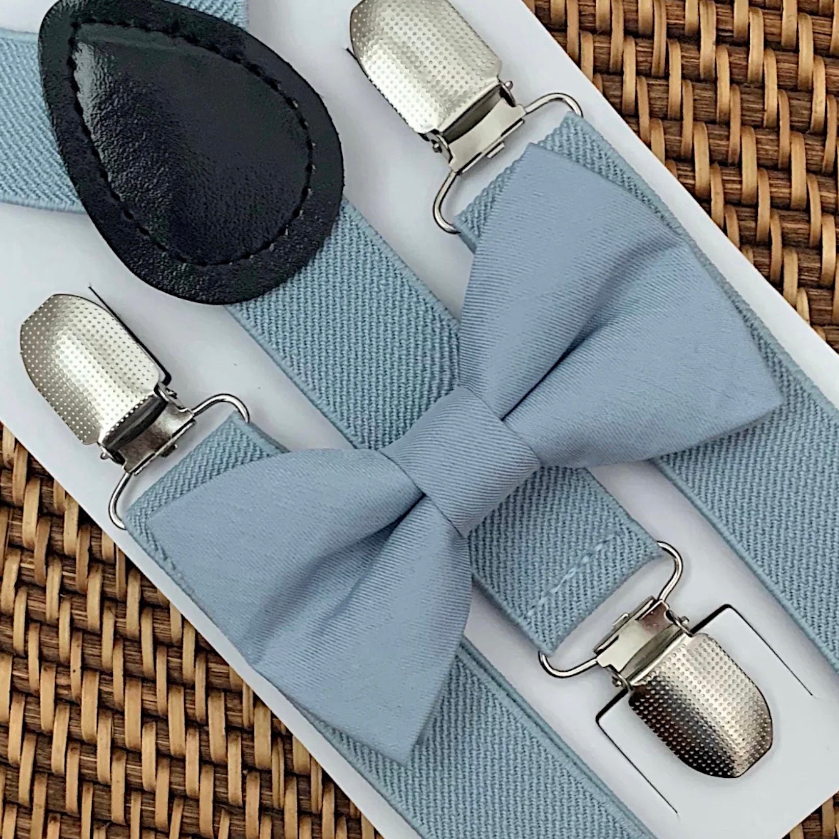 Dusty blue bow tie with silver clips on dusty blue suspenders for groomsmen, ring bearer outfit and wedding on a woven surface