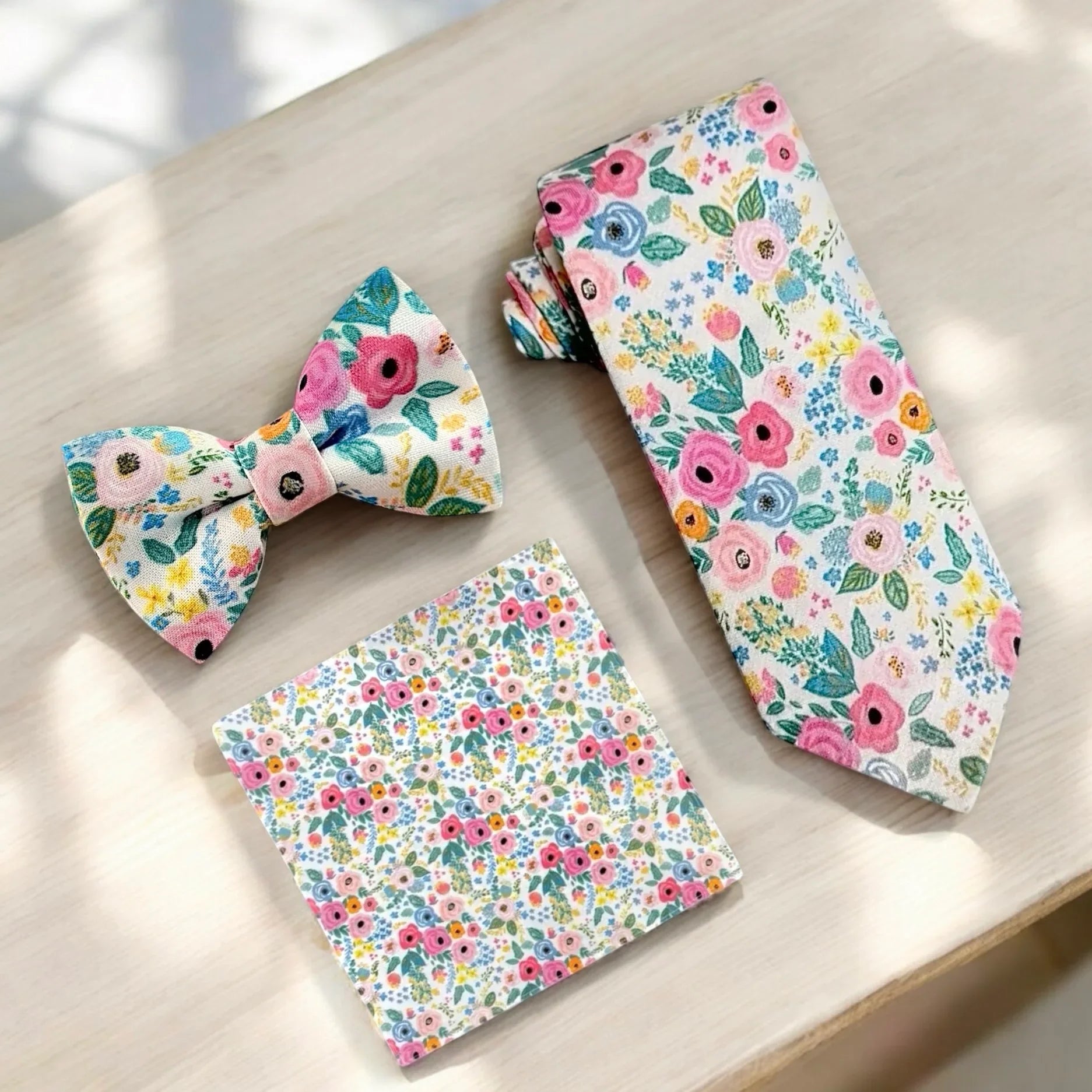 Floral-patterned bow tie, pocket square, and tie on a light wooden surface.