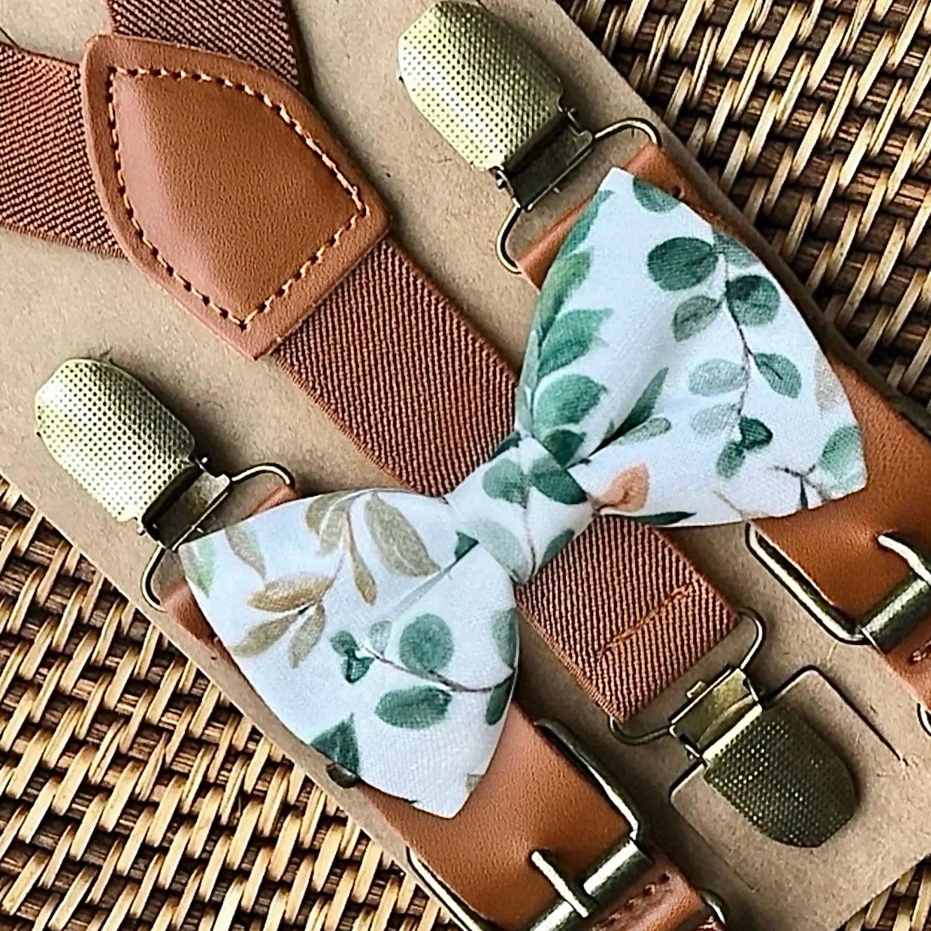 Light blue bow tie with a green leafy pattern paired with cognac leather suspenders and brass buckles on a woven surface.