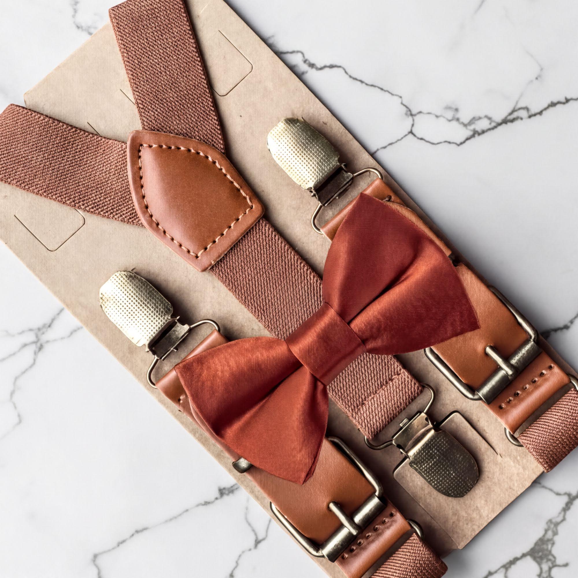 A pair of brown suspenders with a red bow tie attached, displayed on a marble surface.