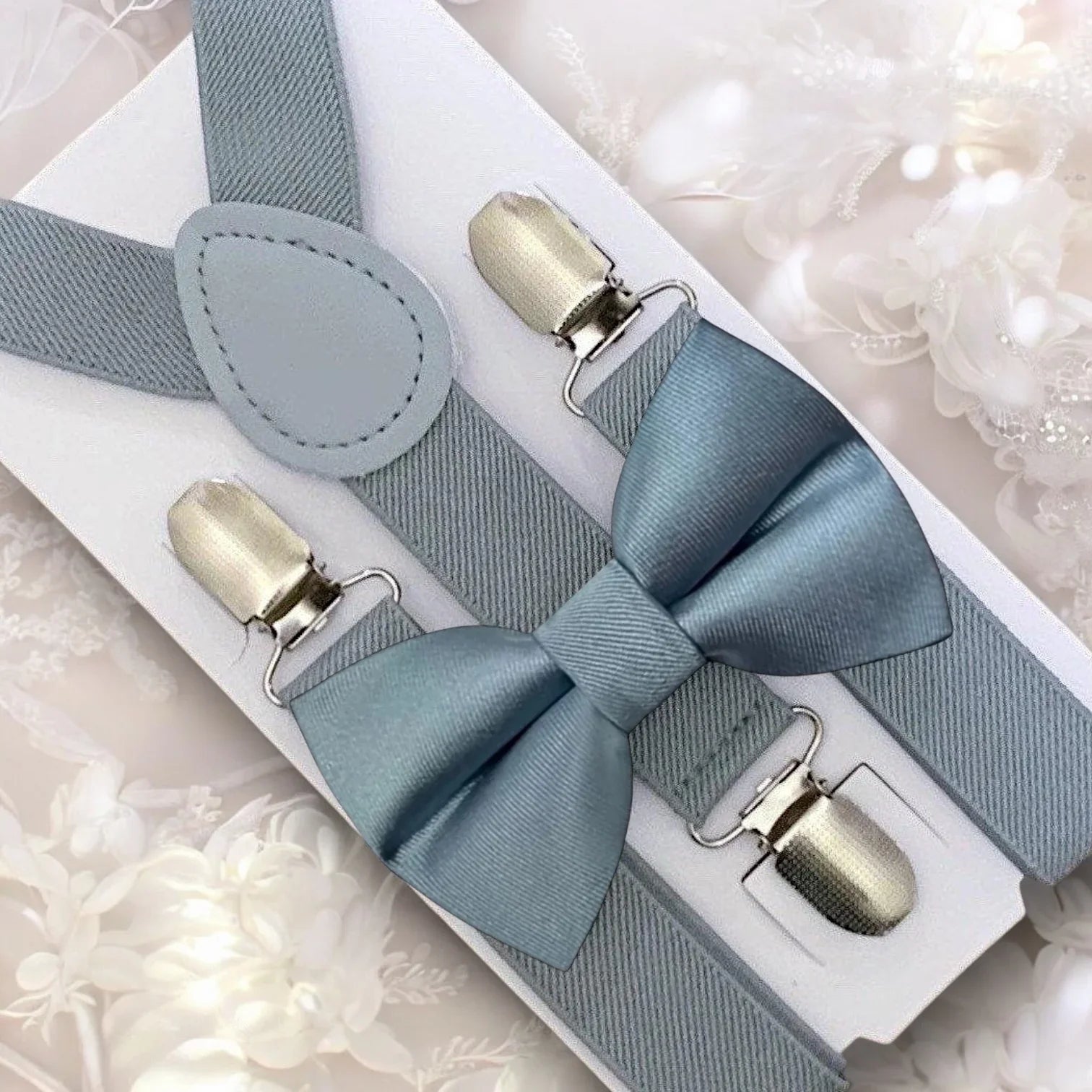Close up of dusty blue bow tie with light gray suspenders arranged on a white display card
