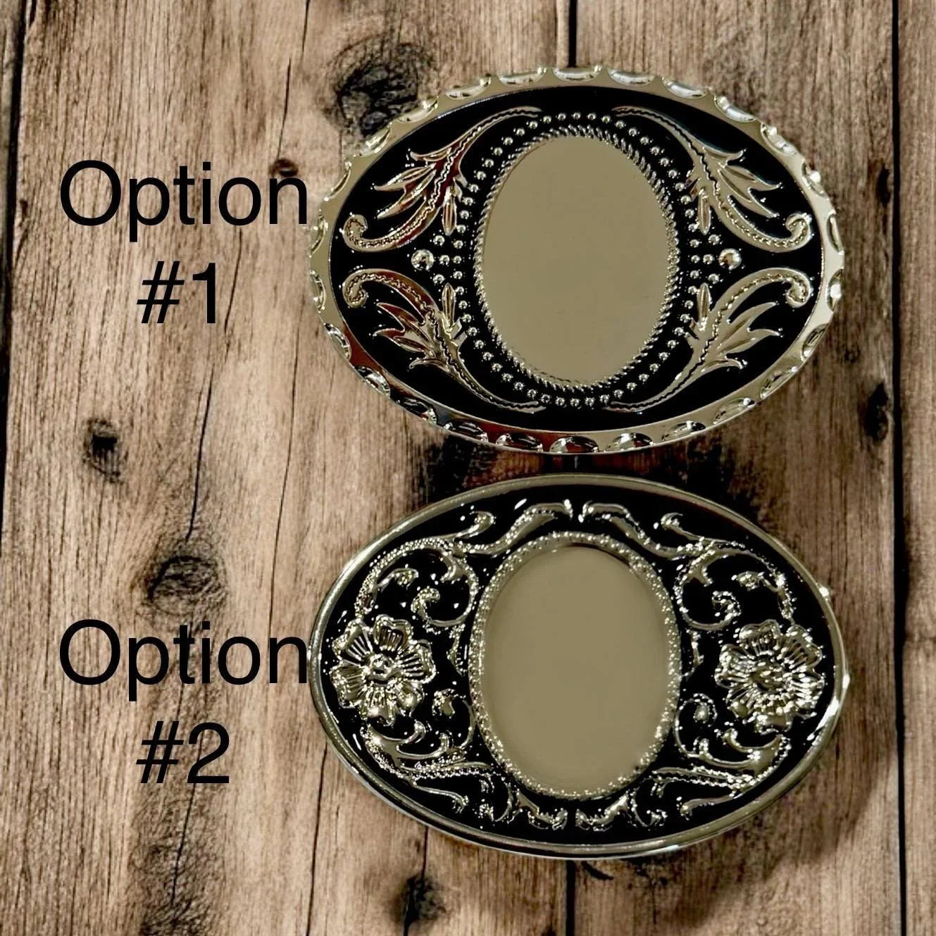 Two oval belt buckles with black enamel and silver scrollwork on a rustic wooden surface