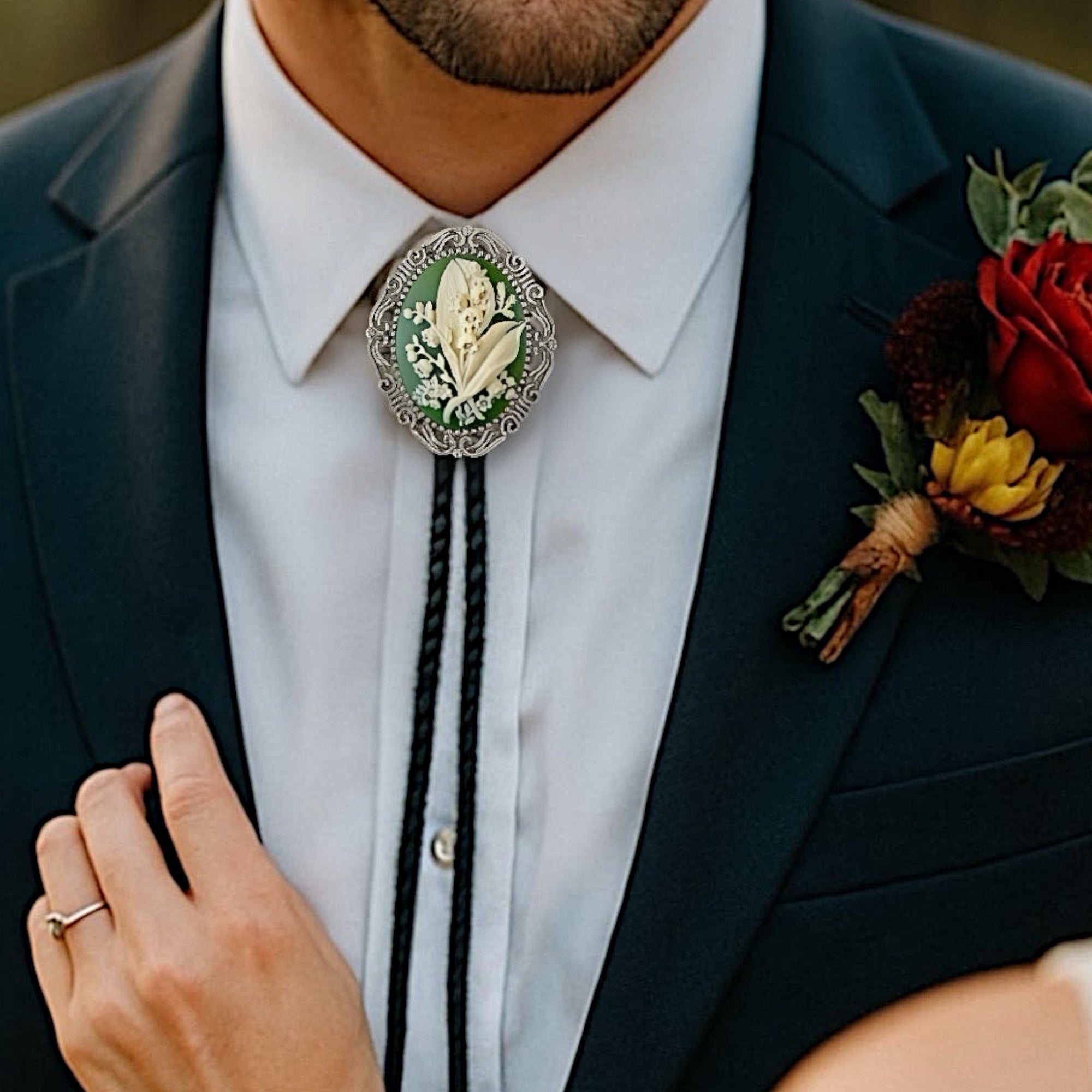 Man wearing a dark suit with a white shirt, decorative May flower green lily of the valley bolo tie, and floral boutonniere.
