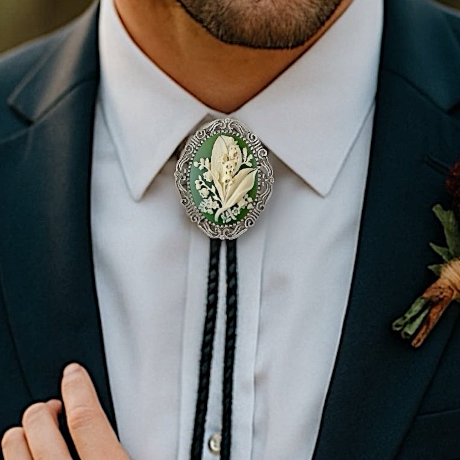 Man wearing a dark suit with a white shirt, decorative May flower green lily of the valley bolo tie, and floral boutonniere.