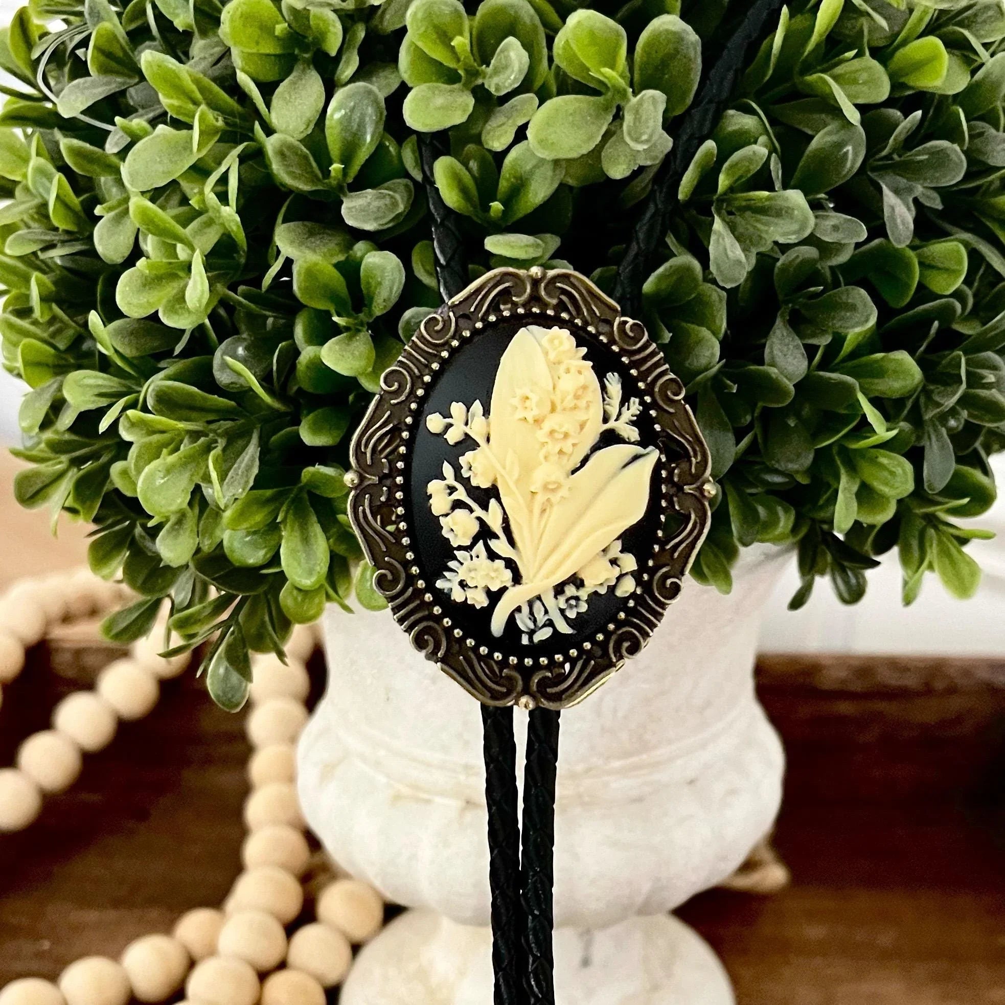Infographic from The Bold Bow Tie titled Bolo Tie Features. Right: A vintage-style bolo tie with a blue and white cowboy riding a horse illustration inside an ornate bronze filigree frame on a black braided cord. Left: Lines connect the image to three checkmark features: Handmade, 39" Long Cord, and Pendant is 1.5" (L) x 1.25" (W). Bottom text reads Perfect Addition to Your Outfit.