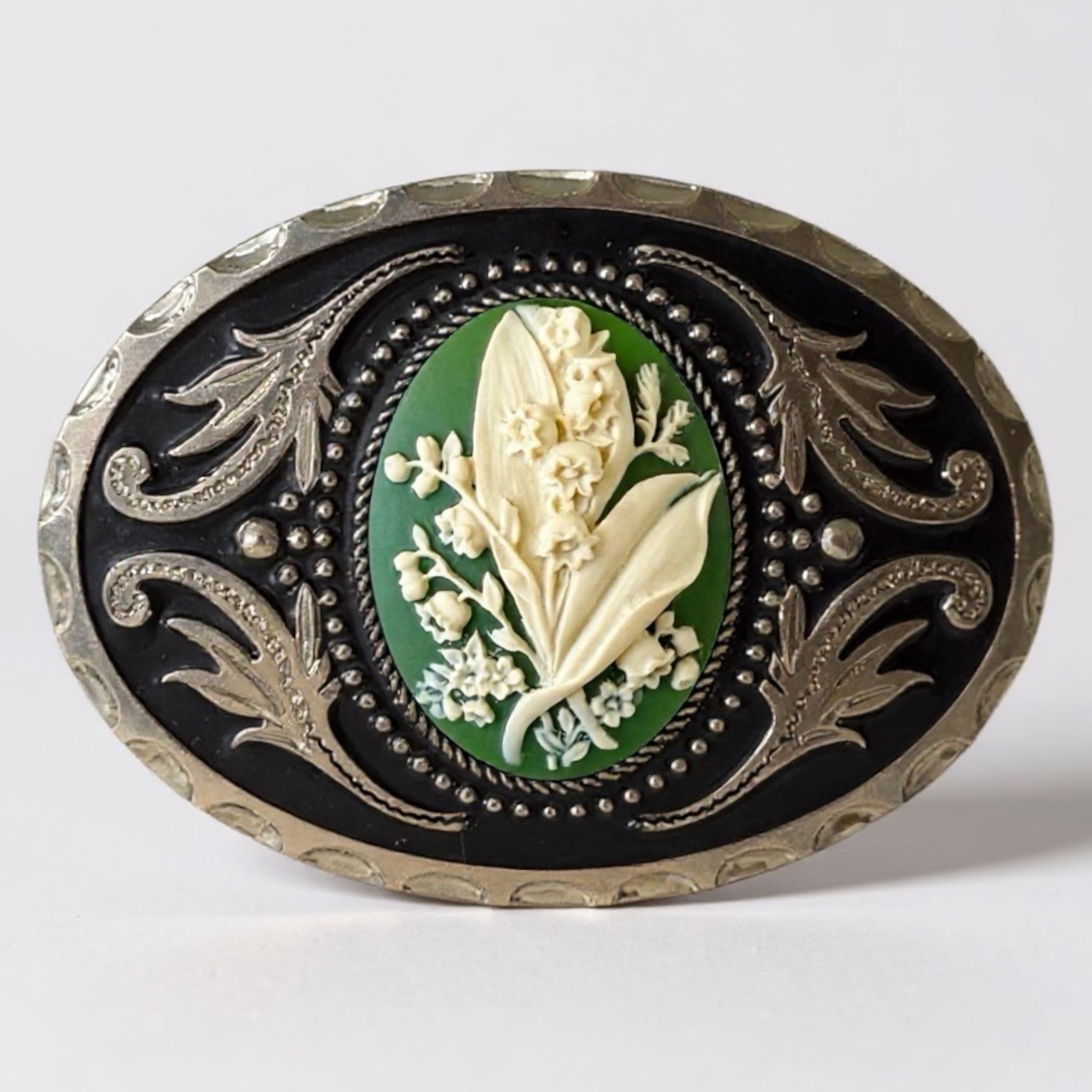 Decorative belt buckle with May flower green lily of the valley floral design and intricate silver border on a light gray background