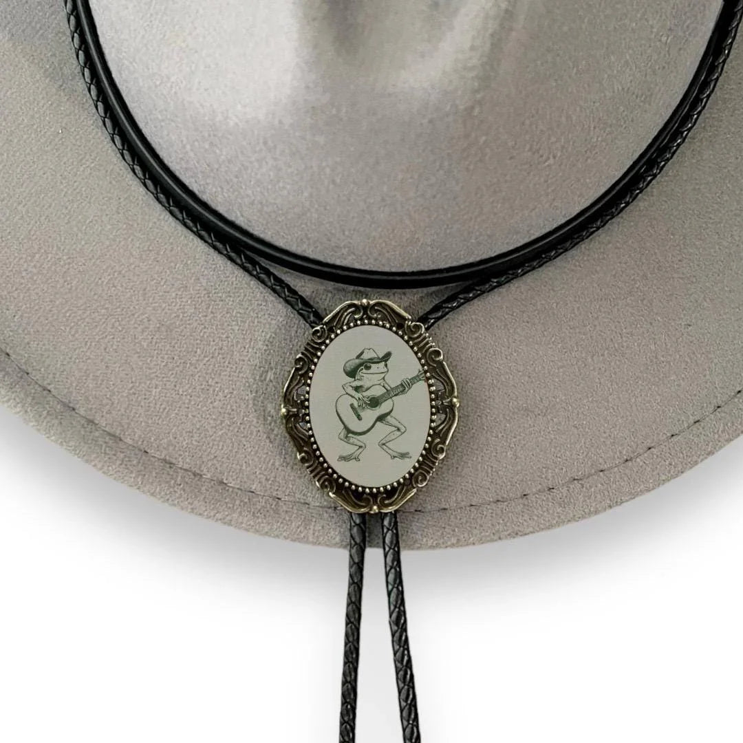 Infographic from The Bold Bow Tie titled Bolo Tie Features. Right: A vintage-style bolo tie with a blue and white cowboy riding a horse illustration inside an ornate bronze filigree frame on a black braided cord. Left: Lines connect the image to three checkmark features: Handmade, 39" Long Cord, and Pendant is 1.5" (L) x 1.25" (W). Bottom text reads Perfect Addition to Your Outfit.