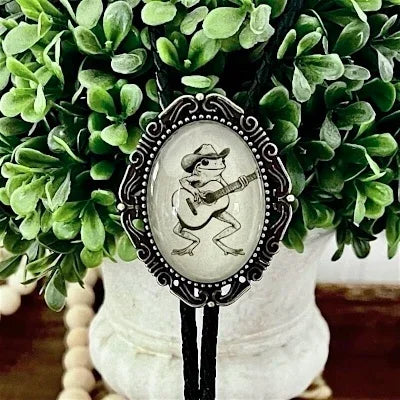 A western-style bolo tie with a black cord and a silver-toned oval pendant featuring a frog playing guitar, displayed in front of green foliage.