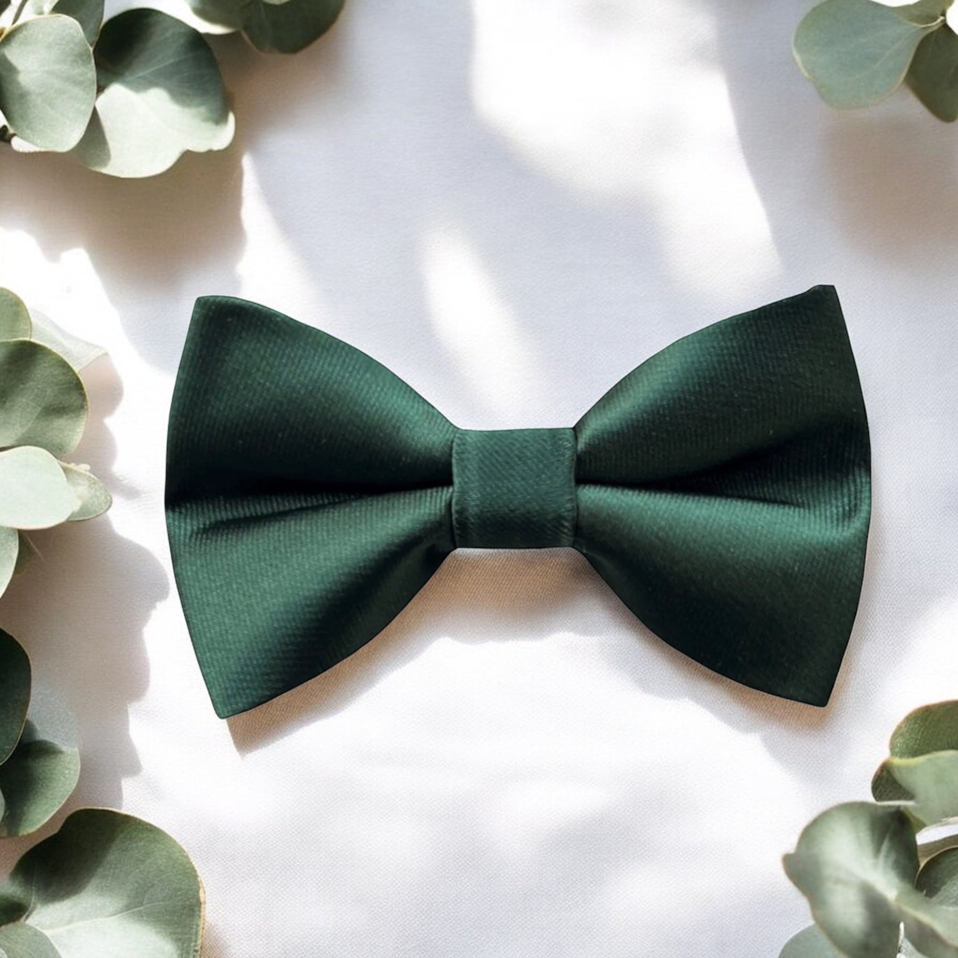 Emerald green bow tie for weddings groomsmen and ring bearers surrounded by eucalyptus on a white tablecloth.