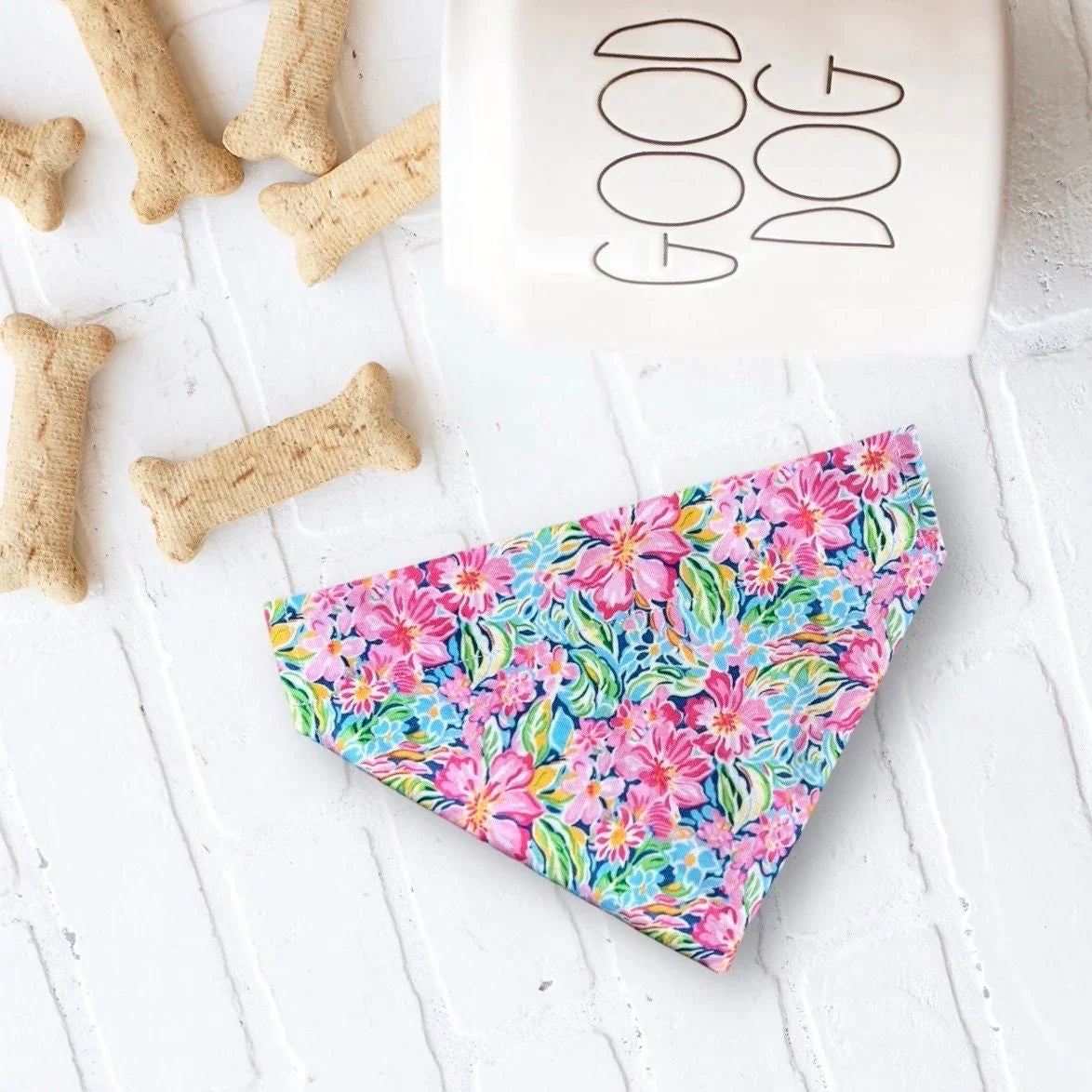Pink floral dog bandana laid on a white textured surface with bone shaped treats and a GOOD DOG sign