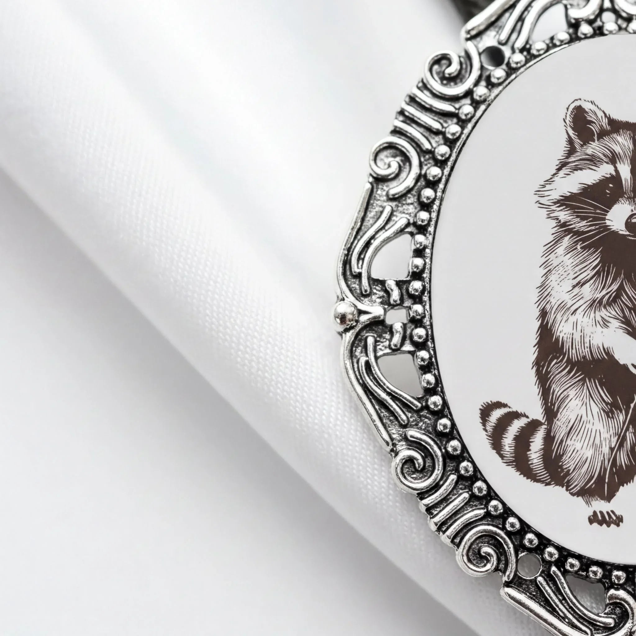 a close-up view of a decorative metal frame with a raccoon illustration on it, placed on a white fabric surface.