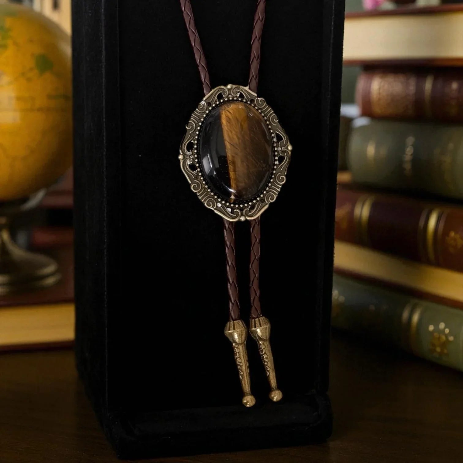 Infographic from The Bold Bow Tie titled Bolo Tie Features. Right: A vintage-style bolo tie with a blue and white cowboy riding a horse illustration inside an ornate bronze filigree frame on a black braided cord. Left: Lines connect the image to three checkmark features: Handmade, 39" Long Cord, and Pendant is 1.5" (L) x 1.25" (W). Bottom text reads Perfect Addition to Your Outfit.