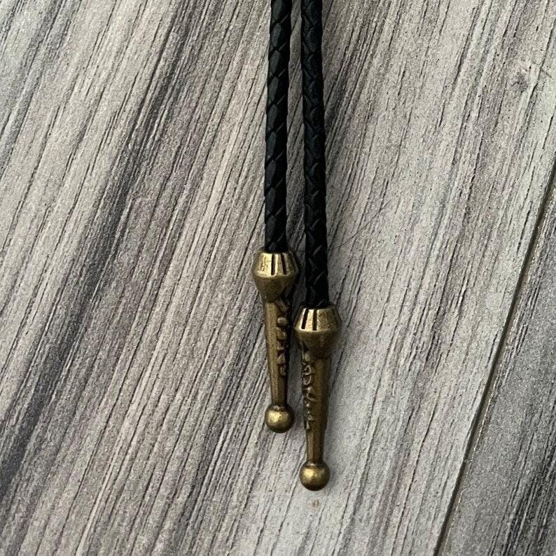 Close-up of Lily of the Valley bolo tie with black braided cords, brass end caps, and a decorative floral pendant on a wood surface