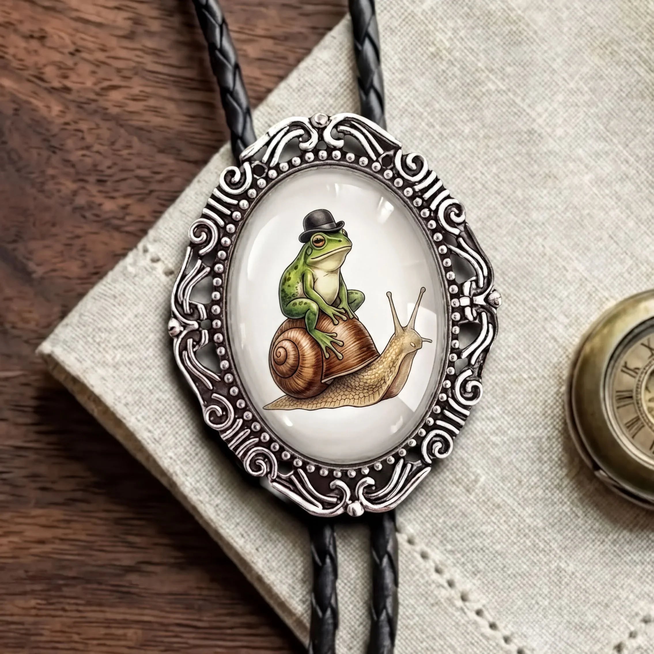A close-up of a bolo tie necklace with a black braided cord and an ornate silver oval pendant. The pendant features an illustration of a green frog sitting on a brown snail wearing a small black hat, set against a light background.