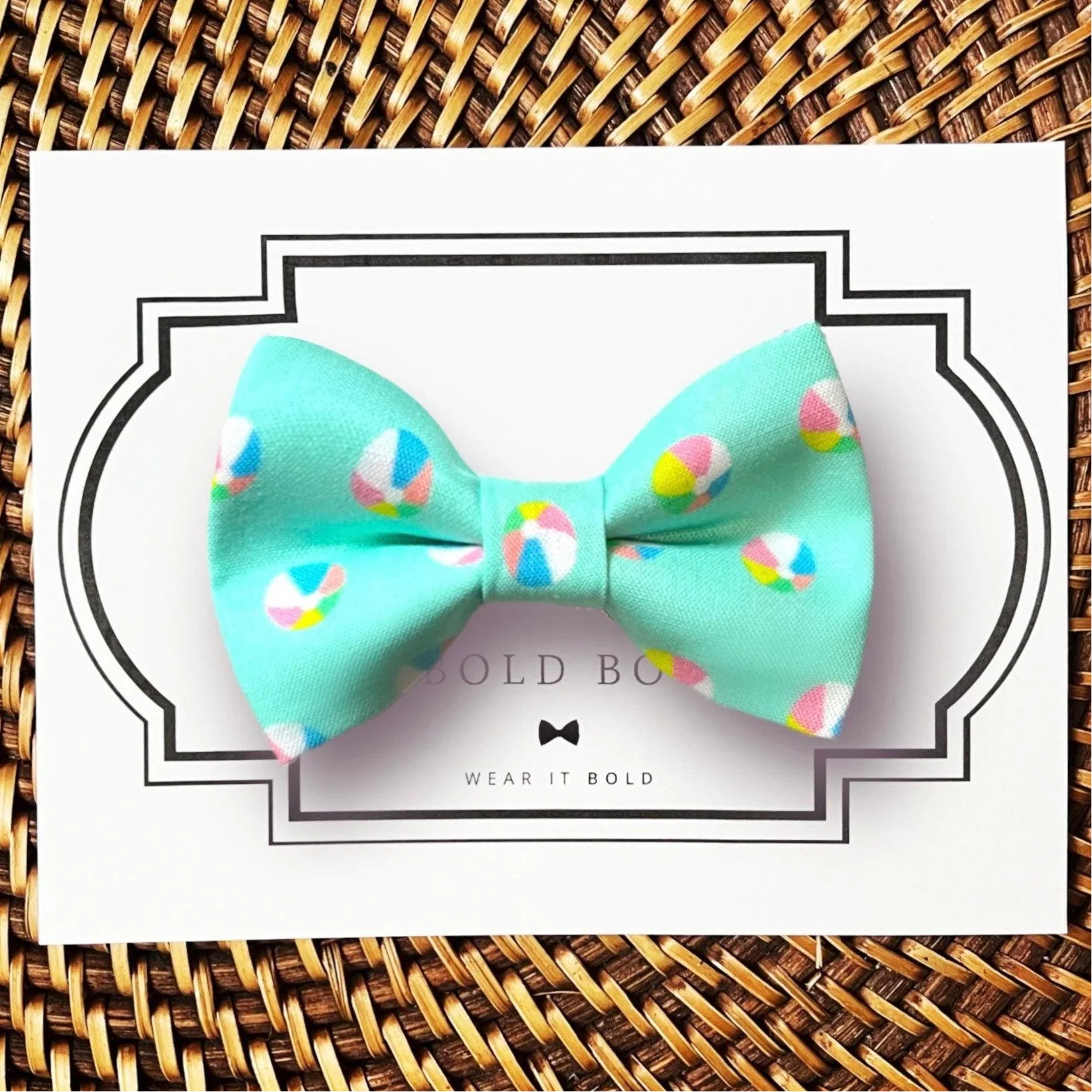 Beachball dog bow tie with colorful patterns on a white card with 'BOLD BOW' branding, placed on a woven surface.
