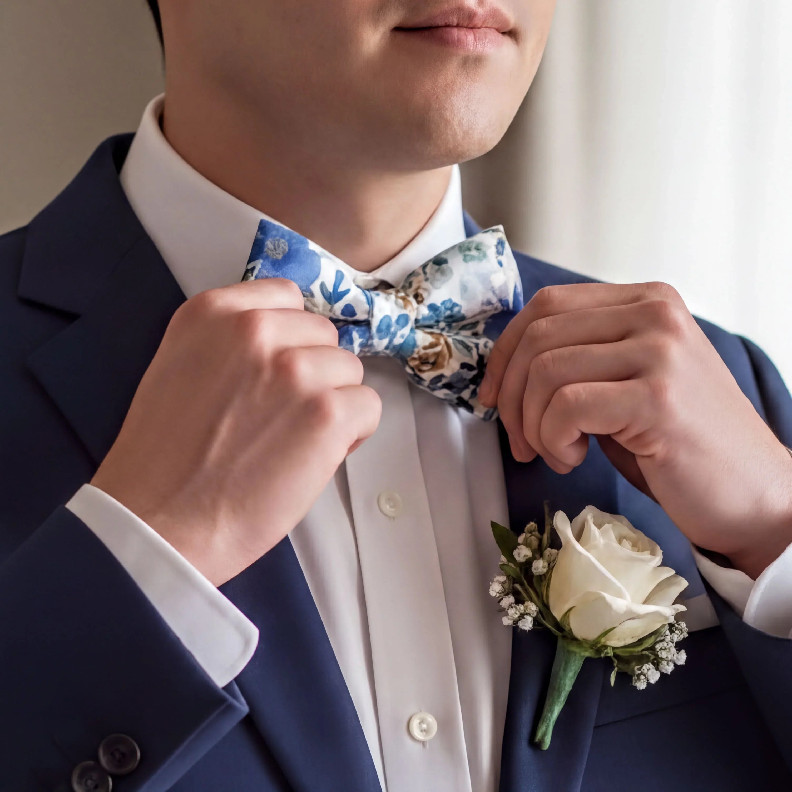 A man in a blue suit jacket and white shirt is adjusting his blue and white floral bowtie.