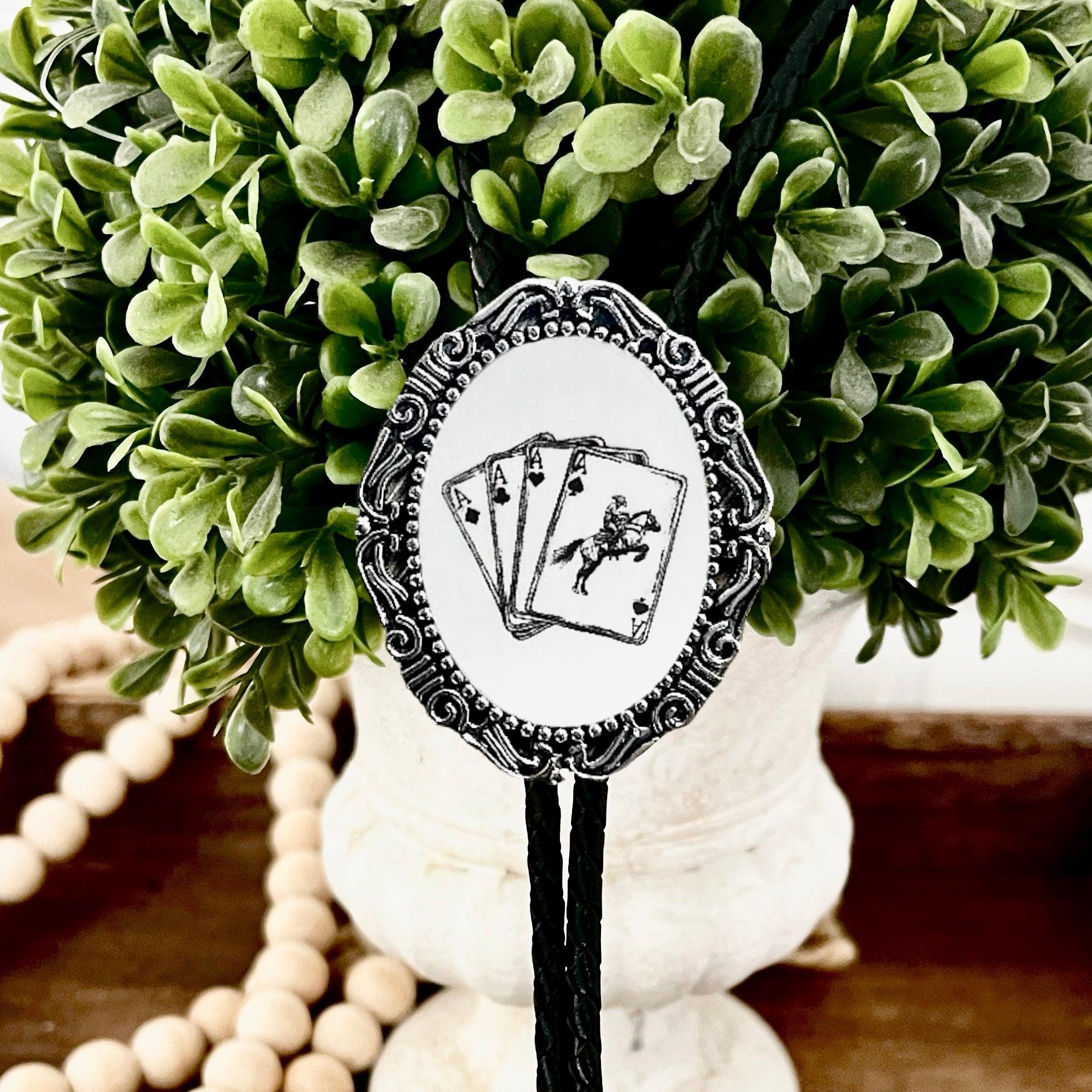 Infographic from The Bold Bow Tie titled Bolo Tie Features. Right: A vintage-style bolo tie with a blue and white cowboy riding a horse illustration inside an ornate bronze filigree frame on a black braided cord. Left: Lines connect the image to three checkmark features: Handmade, 39" Long Cord, and Pendant is 1.5" (L) x 1.25" (W). Bottom text reads Perfect Addition to Your Outfit.