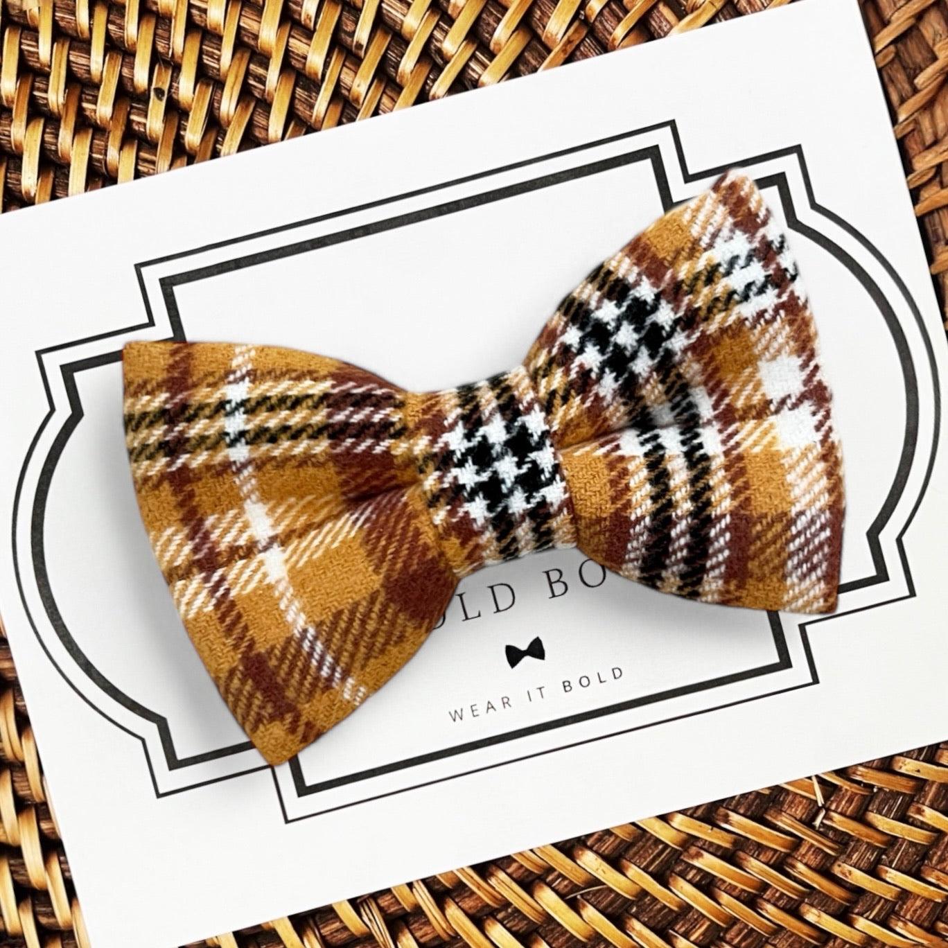 A mustard yellow and gold checkered dog bow tie displayed on a card with 'The Bold Bow Tie' branding.