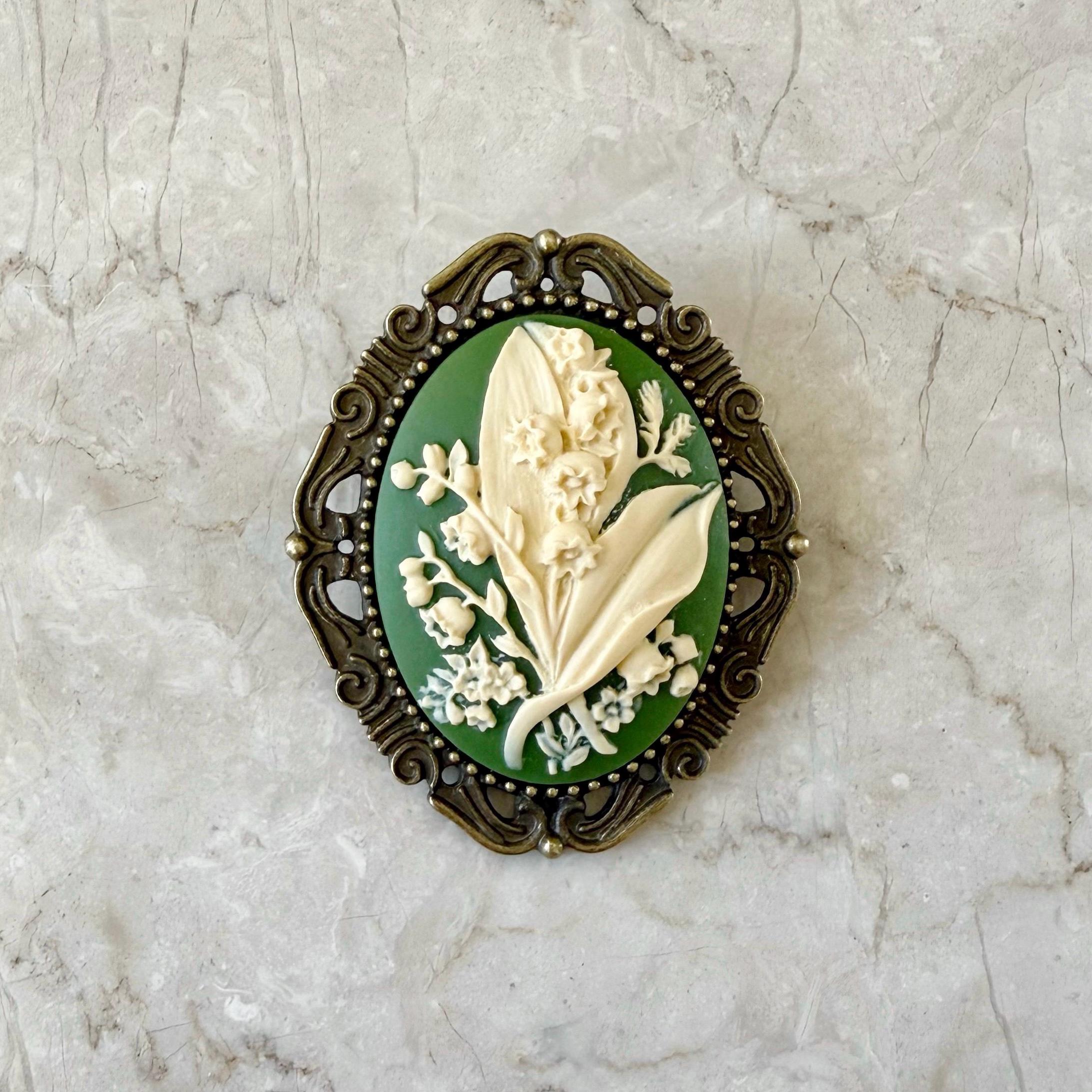 A green and ivory resin cameo brooch with a Lily of the Valley design, set in an antiqued brass frame with a silver pin back.