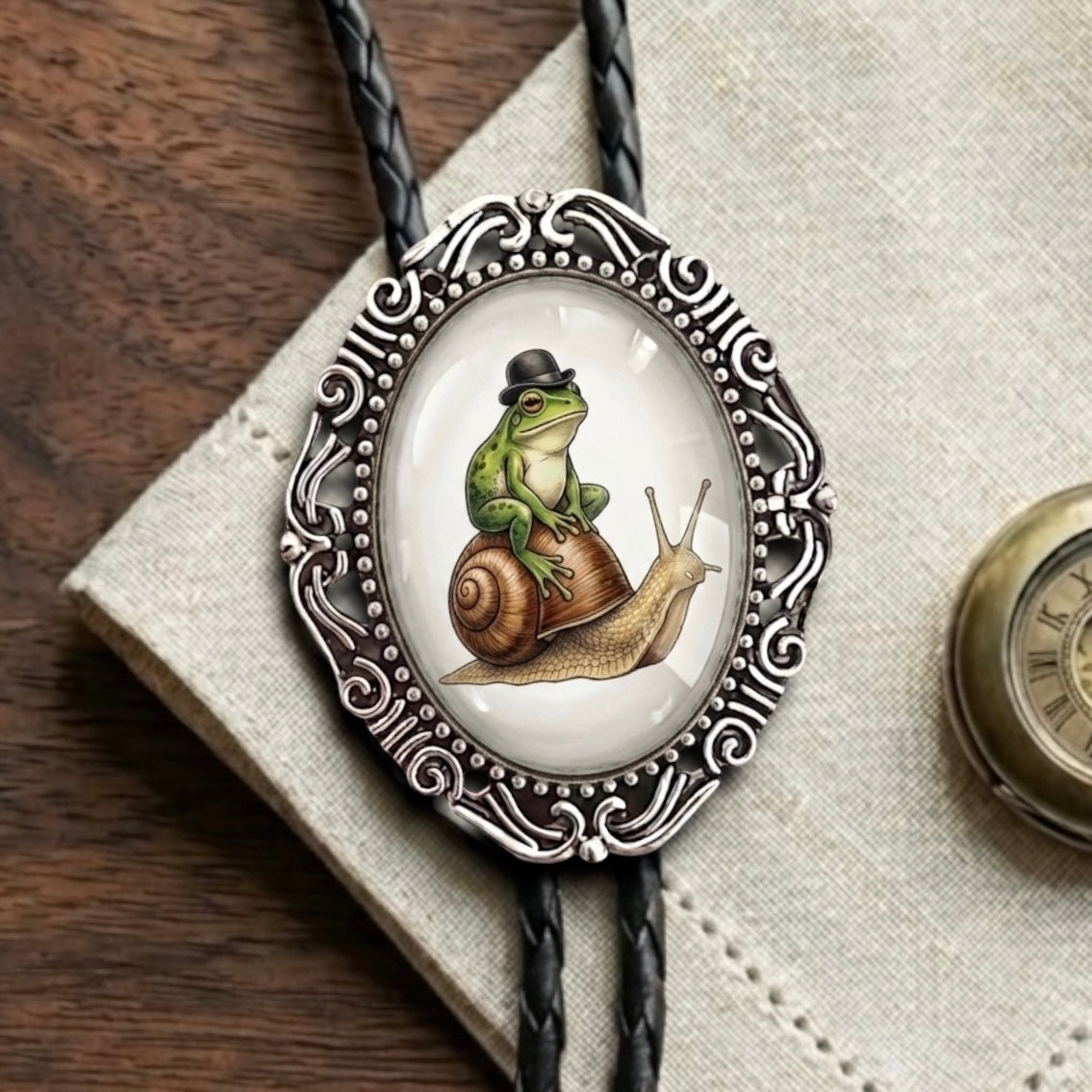 A close-up of a bolo tie necklace with a black braided cord and an ornate silver oval pendant. The pendant features an illustration of a green frog sitting on a brown snail wearing a small black hat, set against a light background.