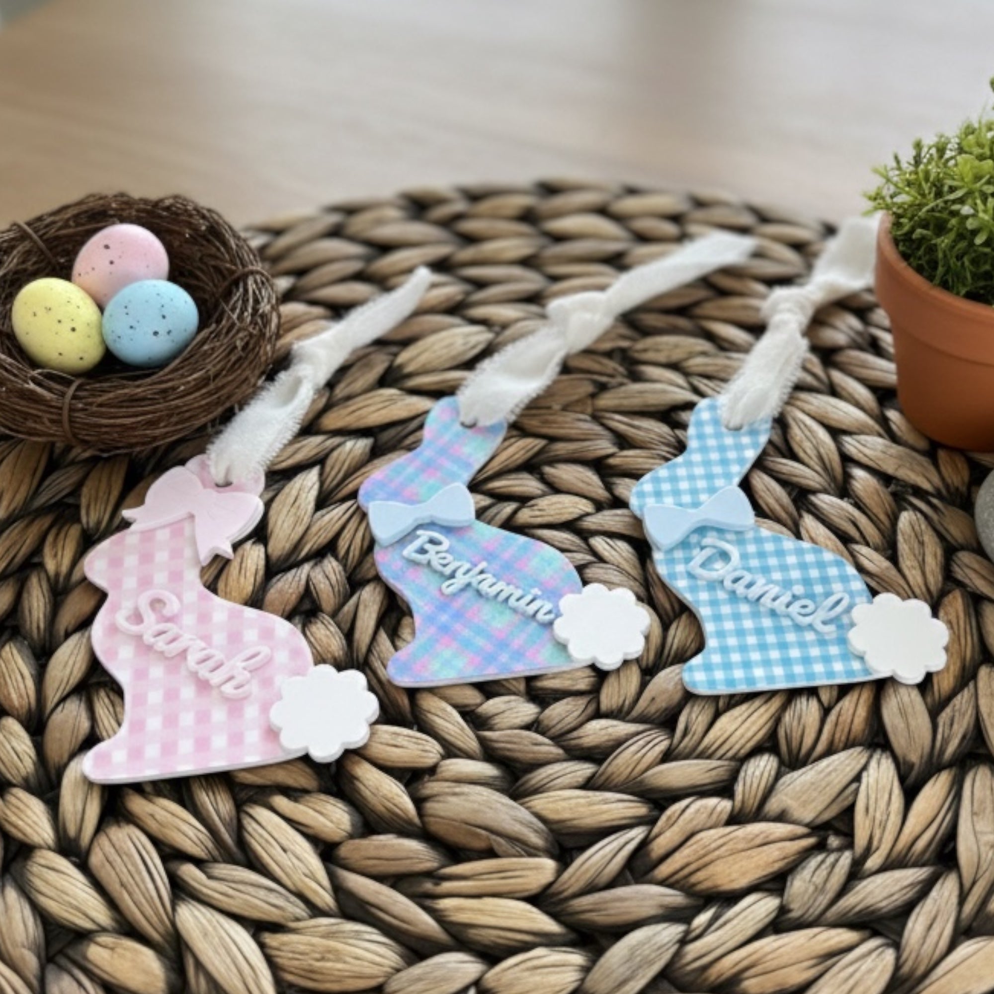 Three decorative Easter bunny tags with names on a woven mat with Easter eggs and a nest.