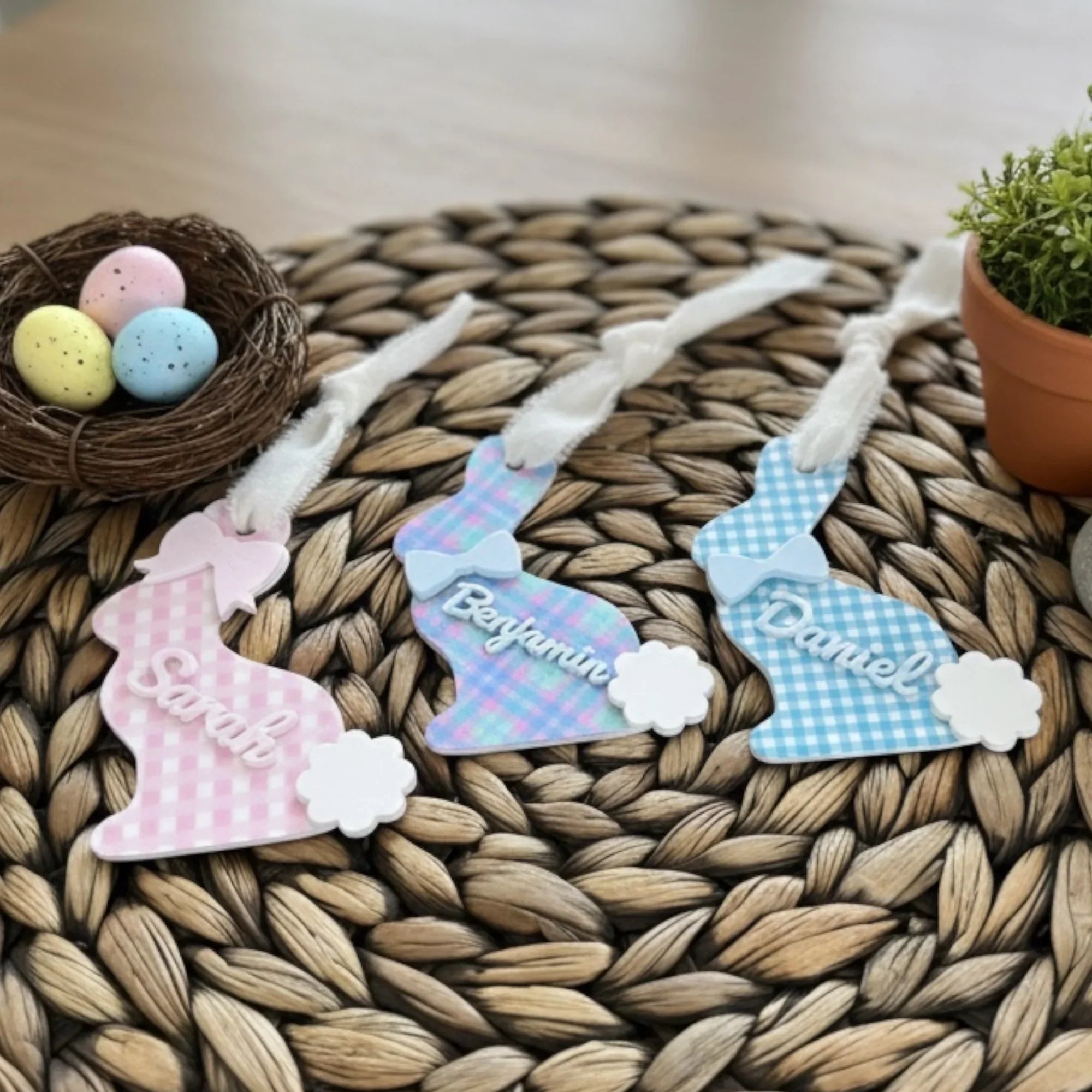 Three decorative Easter bunny tags with names on a woven mat with Easter eggs and a nest.