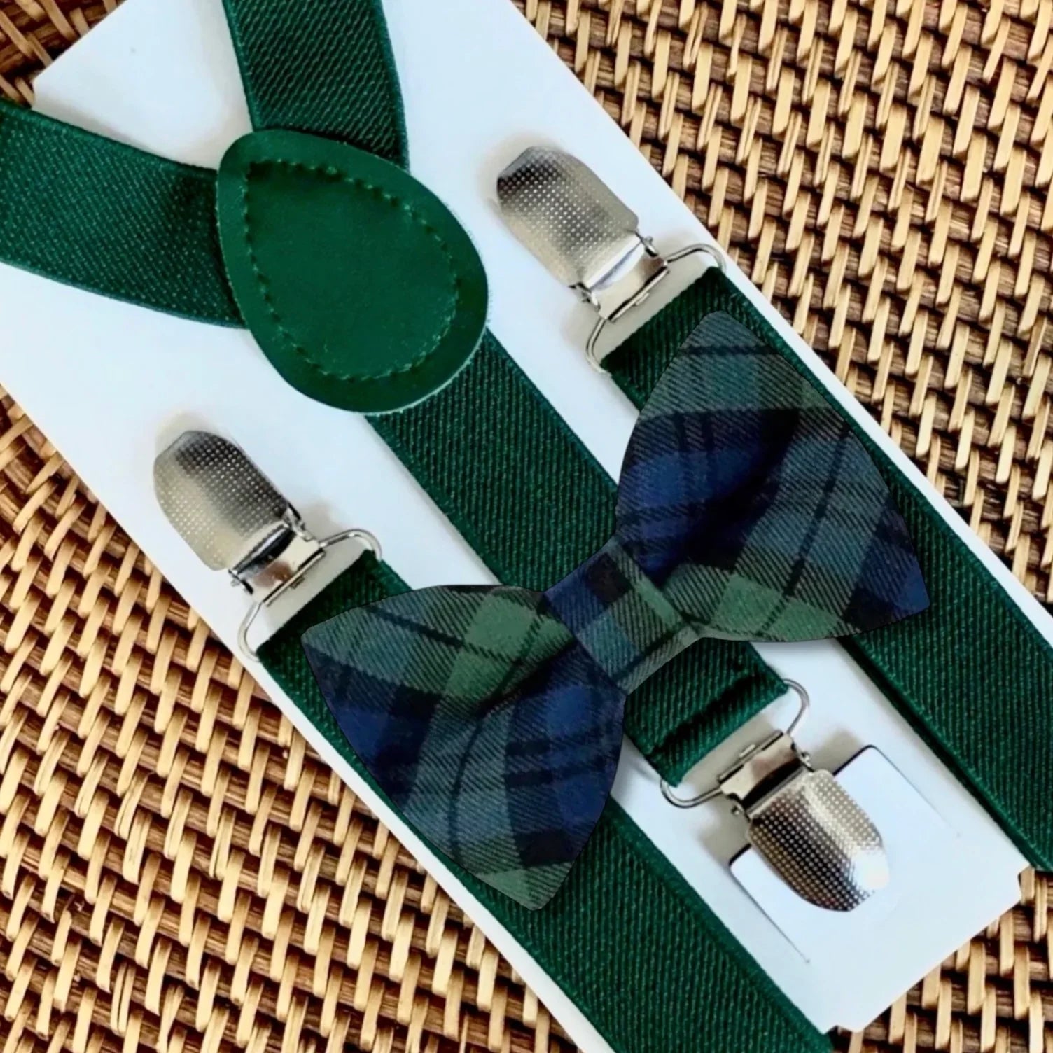 Green plaid bow tie with matching green suspenders arranged on a white card against a wicker background.