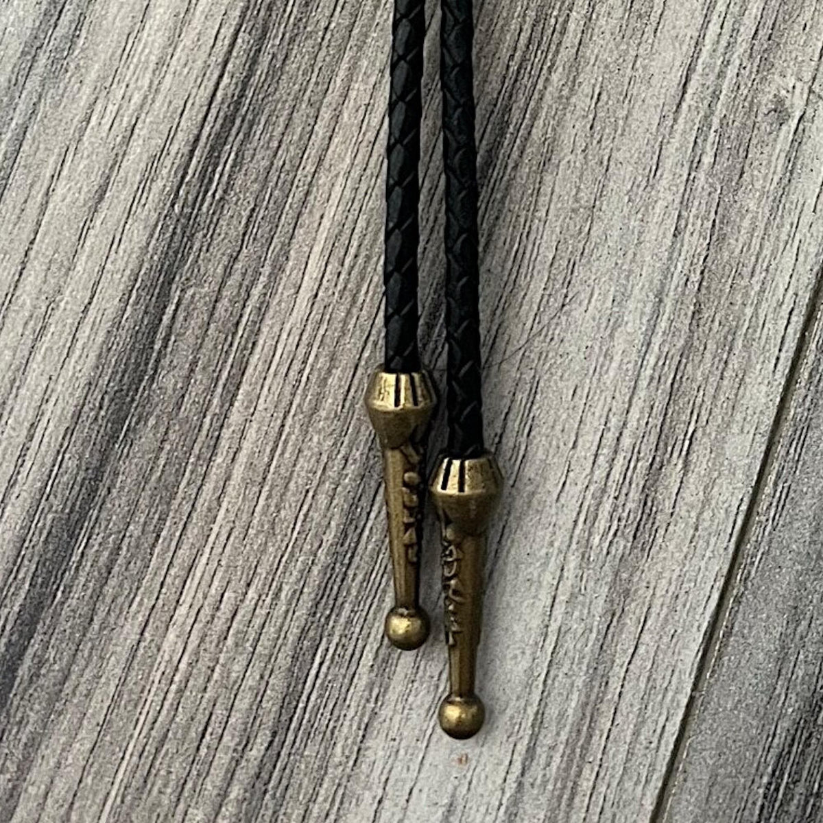 a close-up view of a black braided leather tie with brass-colored metal ends, hanging on a wooden surface.