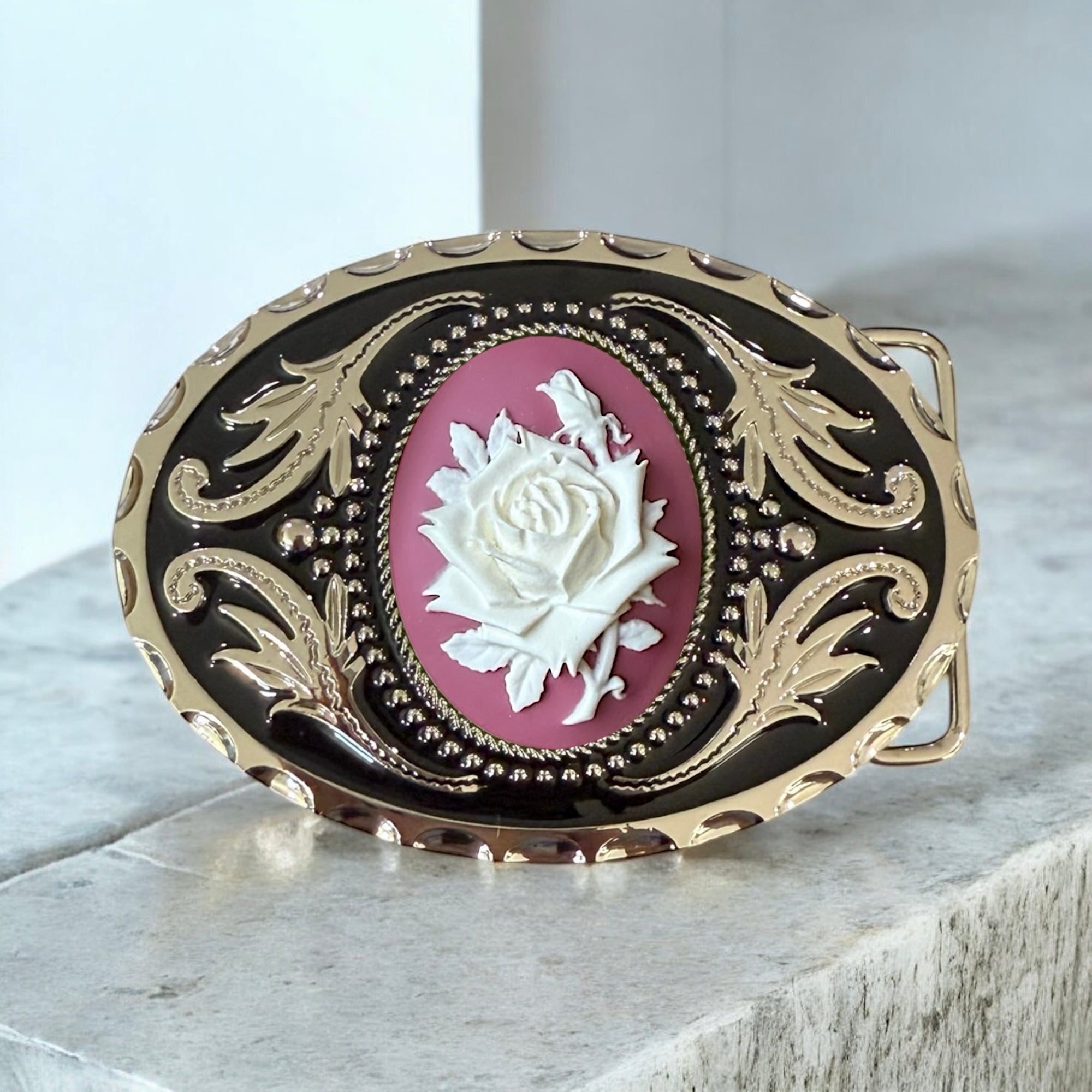 Decorative belt buckle with a white rose design on a pink background, featuring gold and black details.