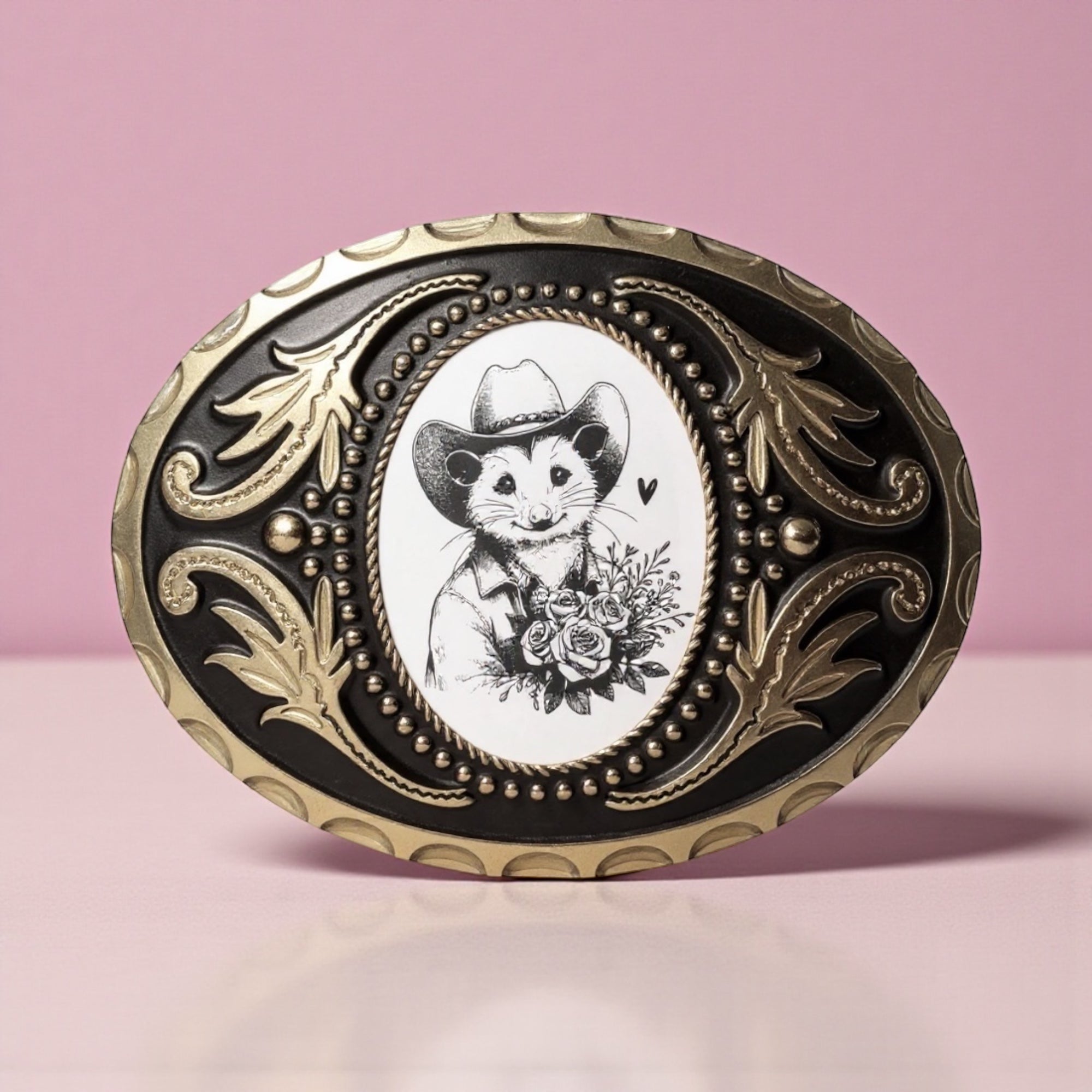 Decorative belt buckle with a opossum wearing a cowboy hat and floral design on a pink background