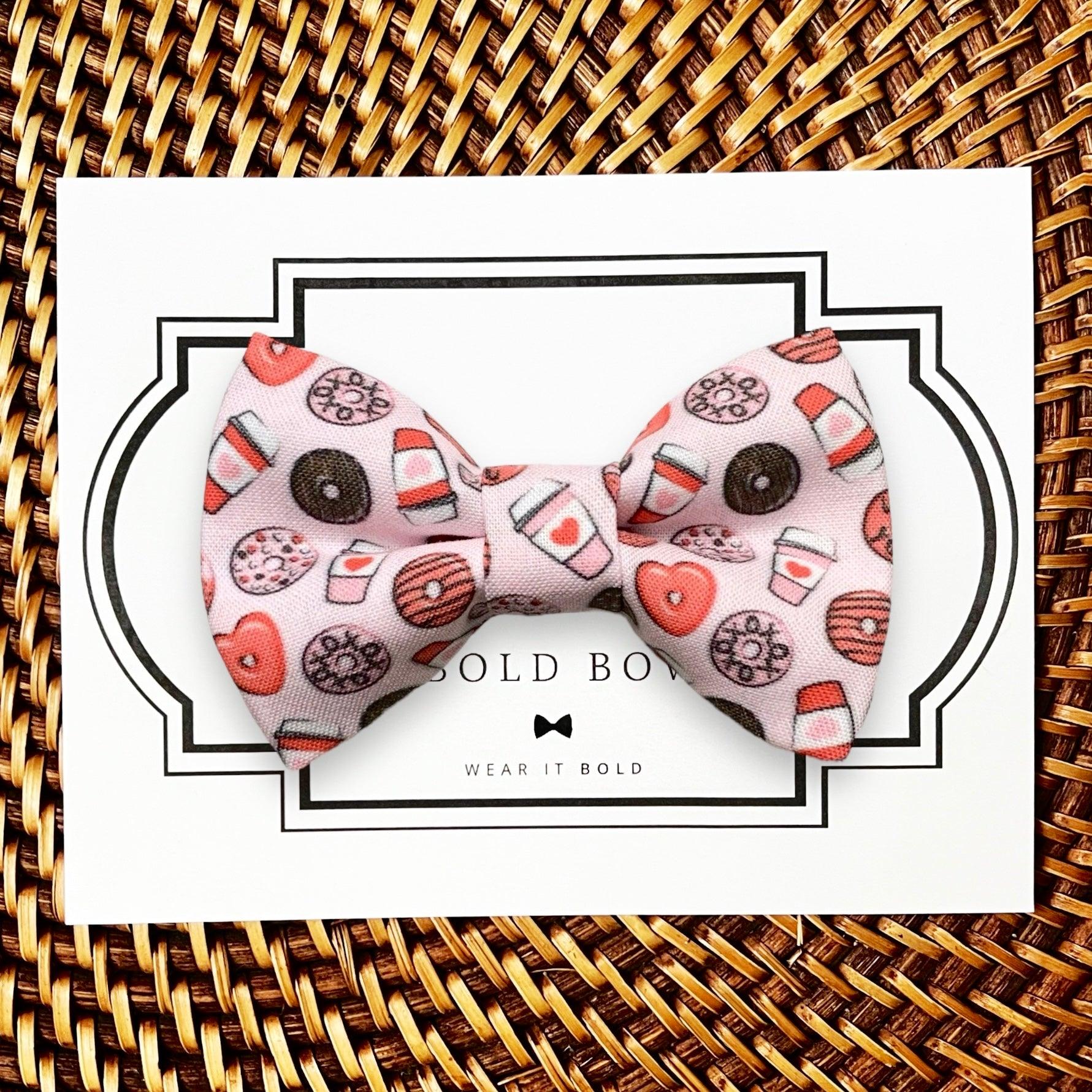 Pink dog bow tie with coffee and donut pattern on a white card with 'The Bold Bow Tie, Wear It Bold' branding, placed on a woven surface.