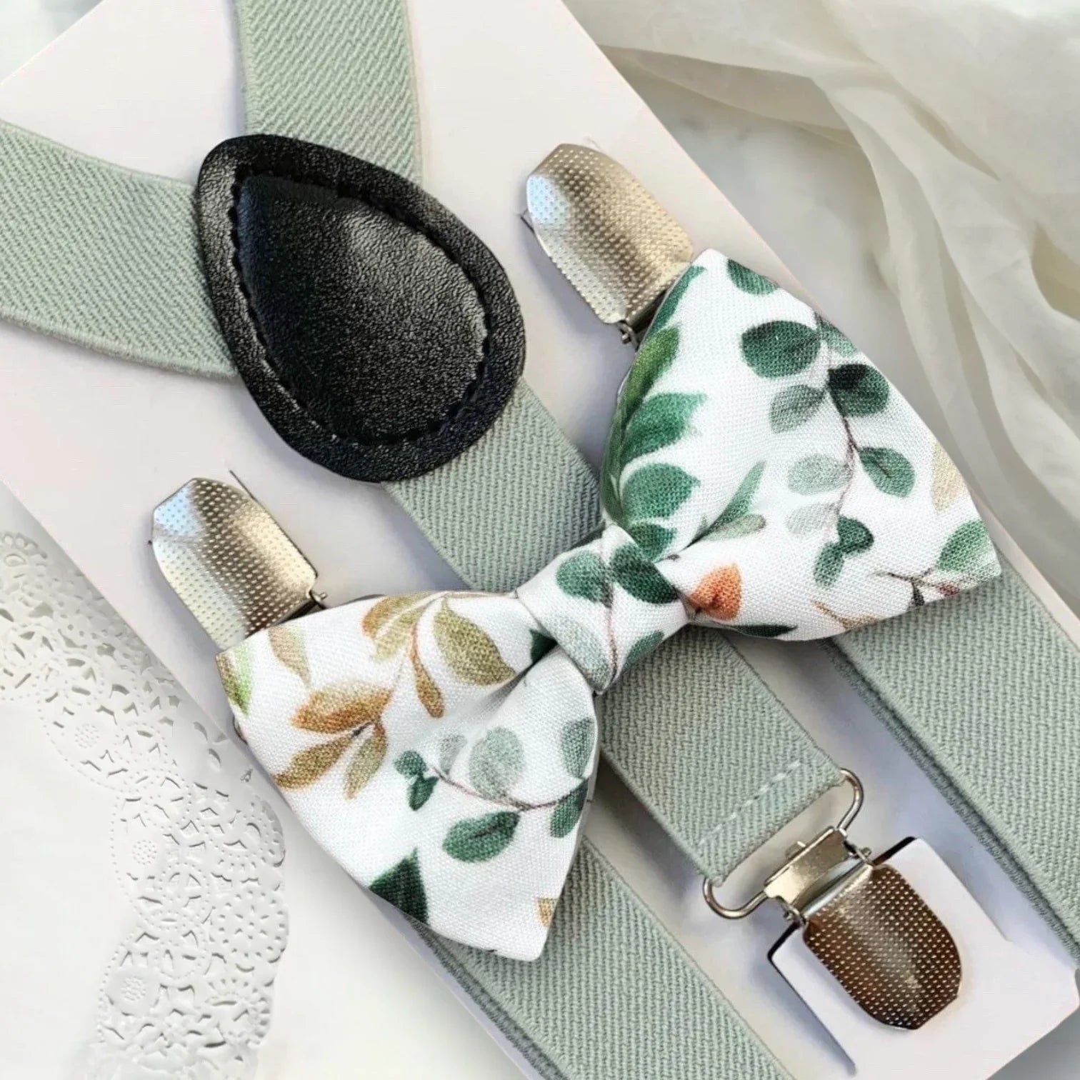 Flat lay of Floral Sage Bow Tie and matching sage suspenders with metal clips on a white background.