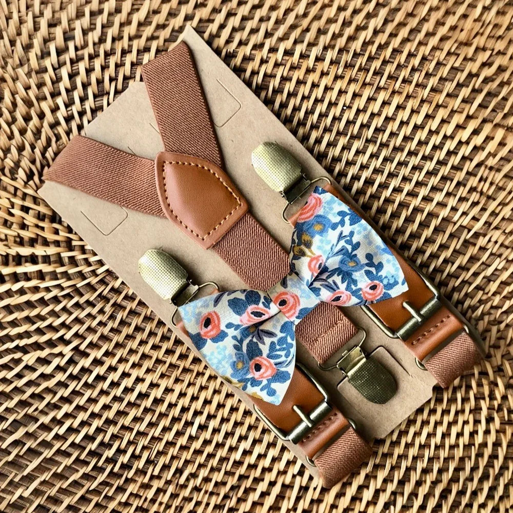Close up of white floral bow tie with cognac brown leather suspenders on a woven basket background
