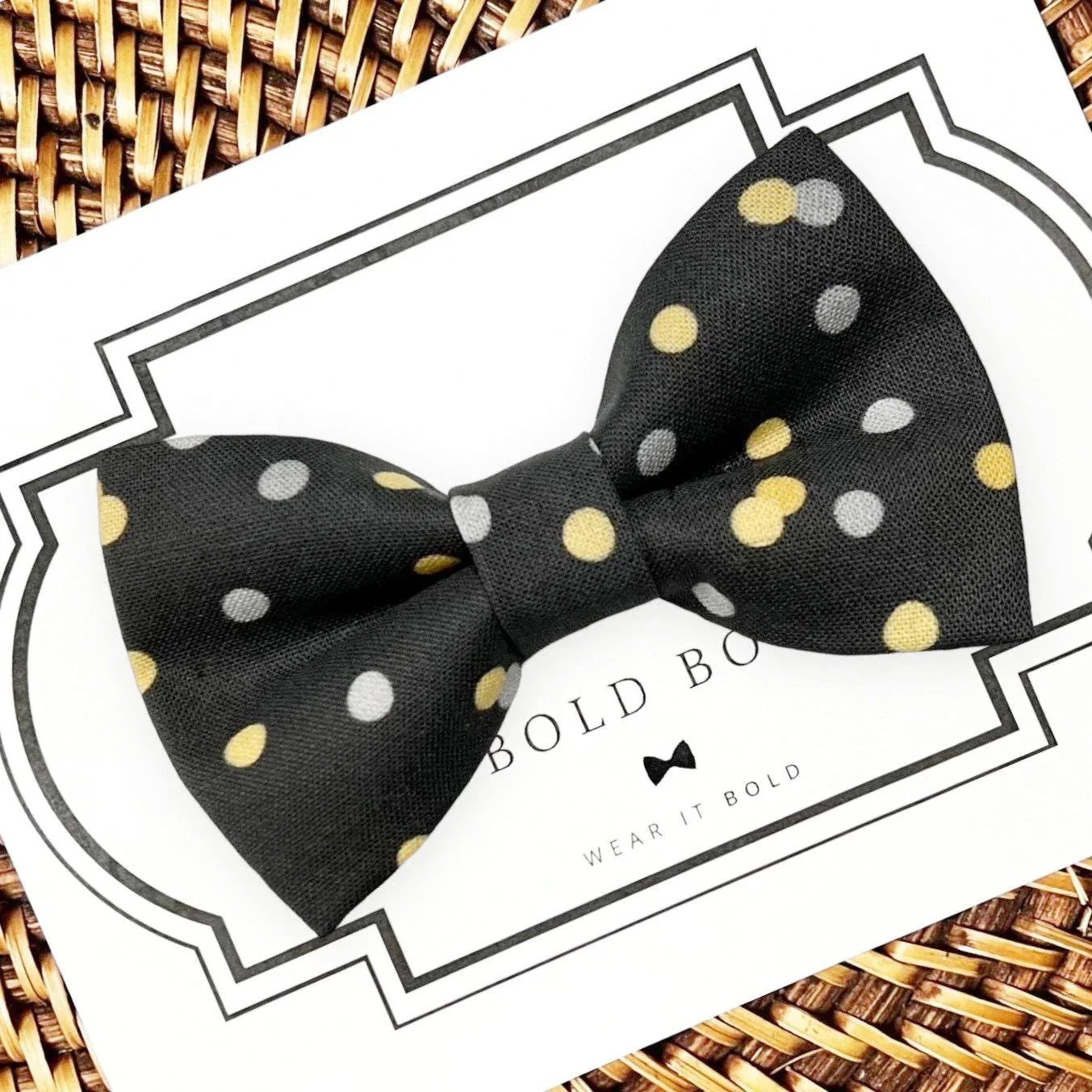 Black polka dot bow tie with yellow and gray dots on a white display card atop a woven basket background.