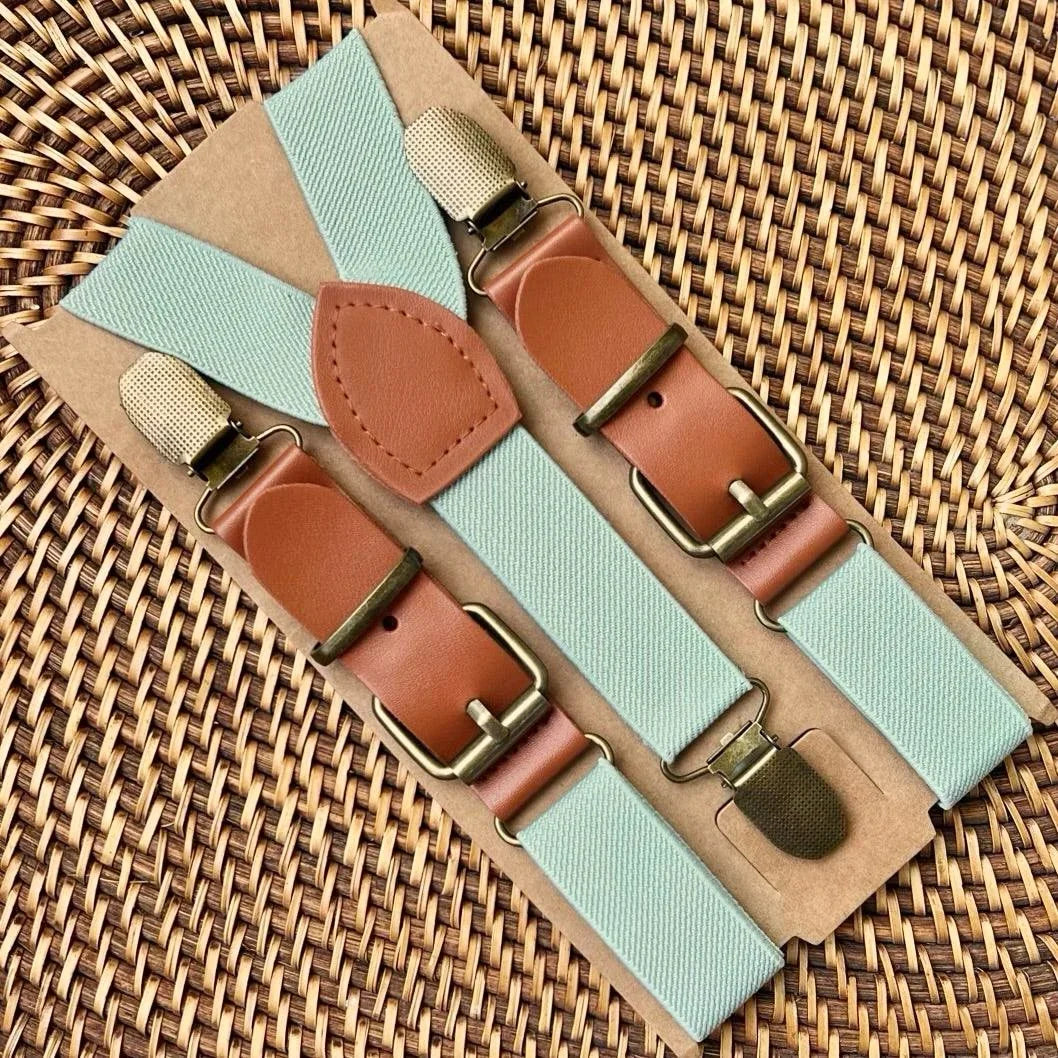 Sage green suspenders with tan leather patches and brass hardware laid on a woven background for weddings