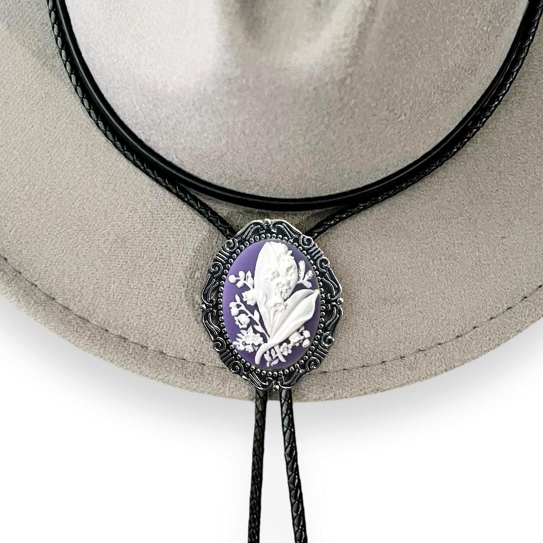 Purple Lily of the Valley bolo tie with white relief flowers on a purple cameo and decorative metal frame on black cords.