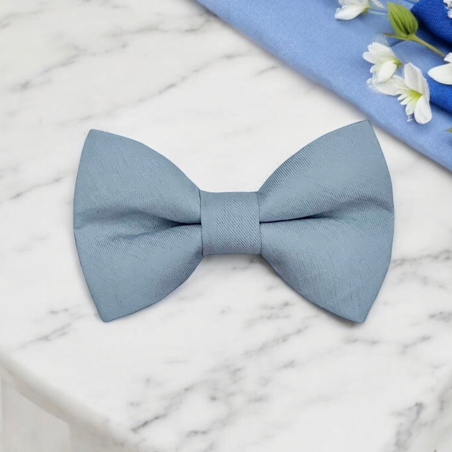 Dusty blue bow tie on a marble surface with a blue cloth and flowers in the background